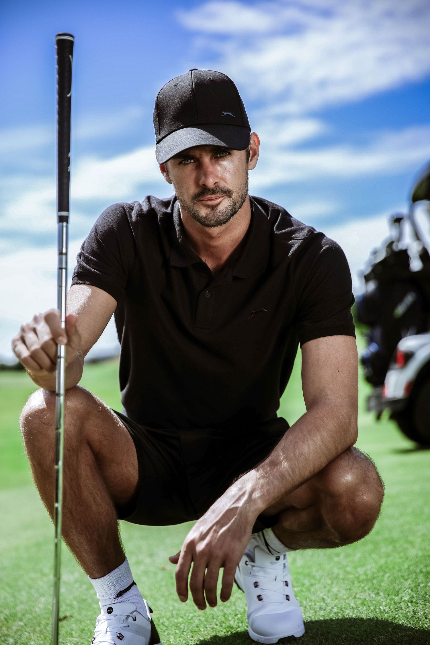 Male golfer in black polo and cap crouches on green holding golf club under blue sky.