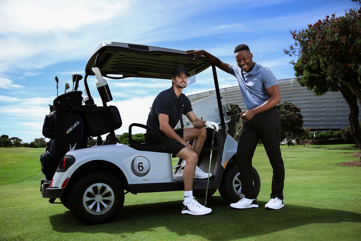 Two men in golf attire pose casually beside a white golf cart on a course.