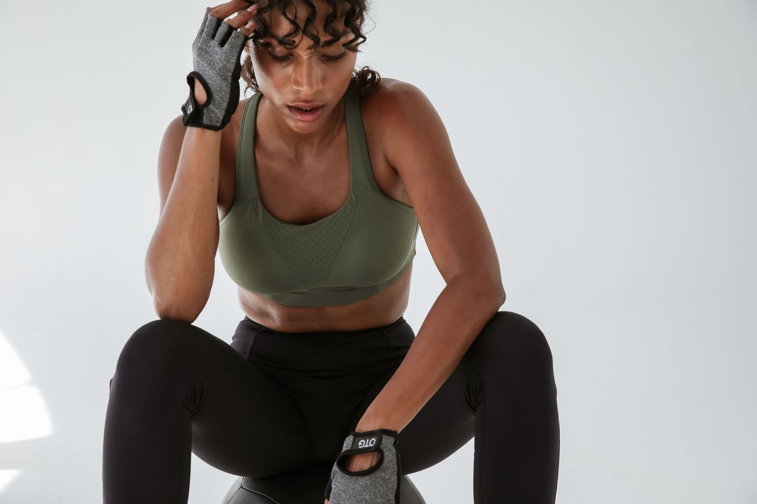 Woman in olive sports bra and black leggings wearing workout gloves resting during fitness training session.