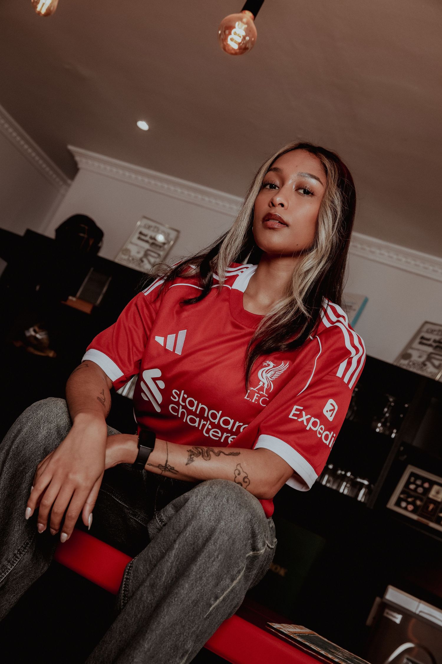 Woman wearing a red Liverpool FC Adidas jersey sitting casually in a dimly lit room.
