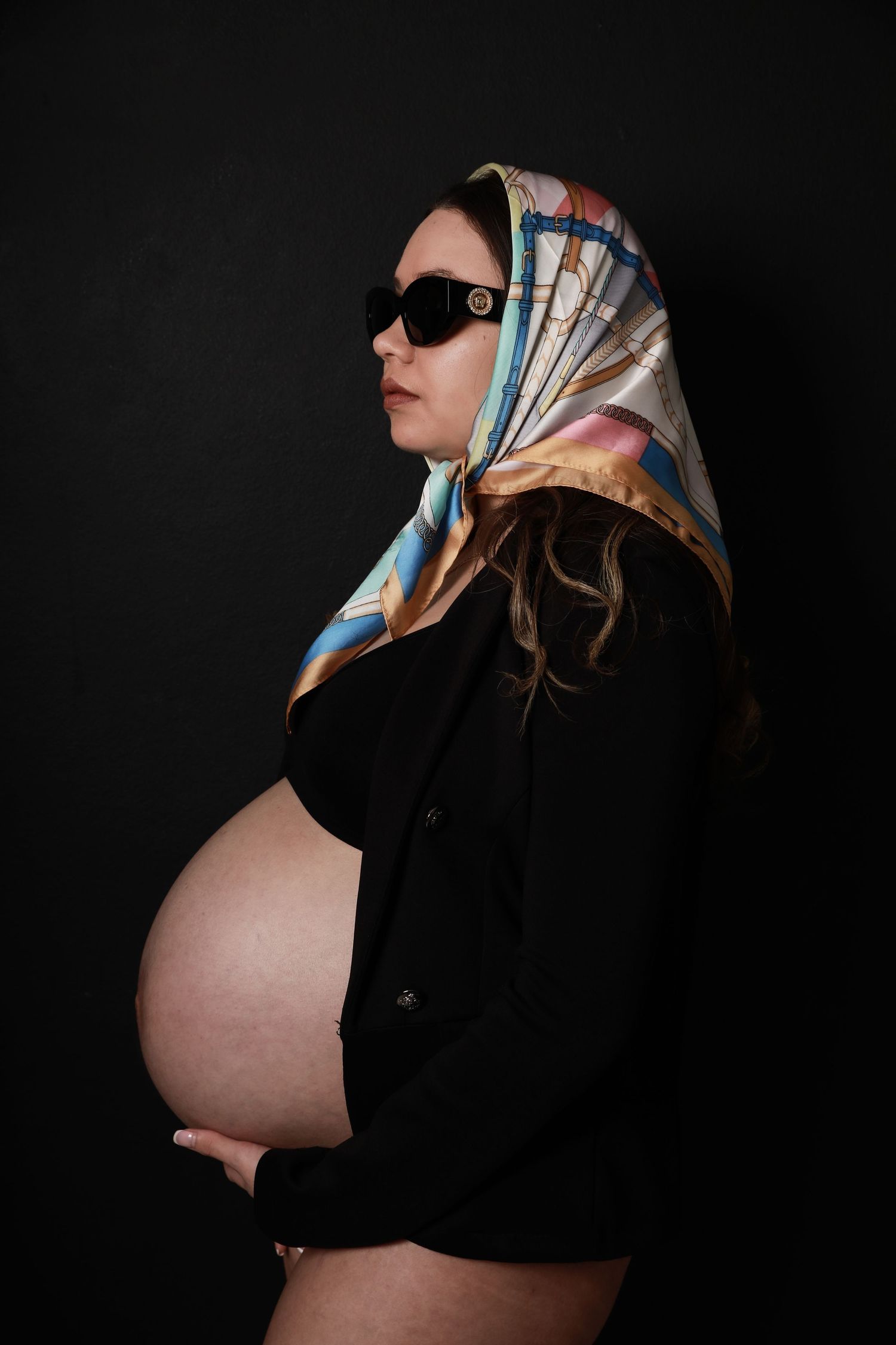 A pregnant woman poses in profile wearing sunglasses and a colorful silk headscarf against a dark background.
