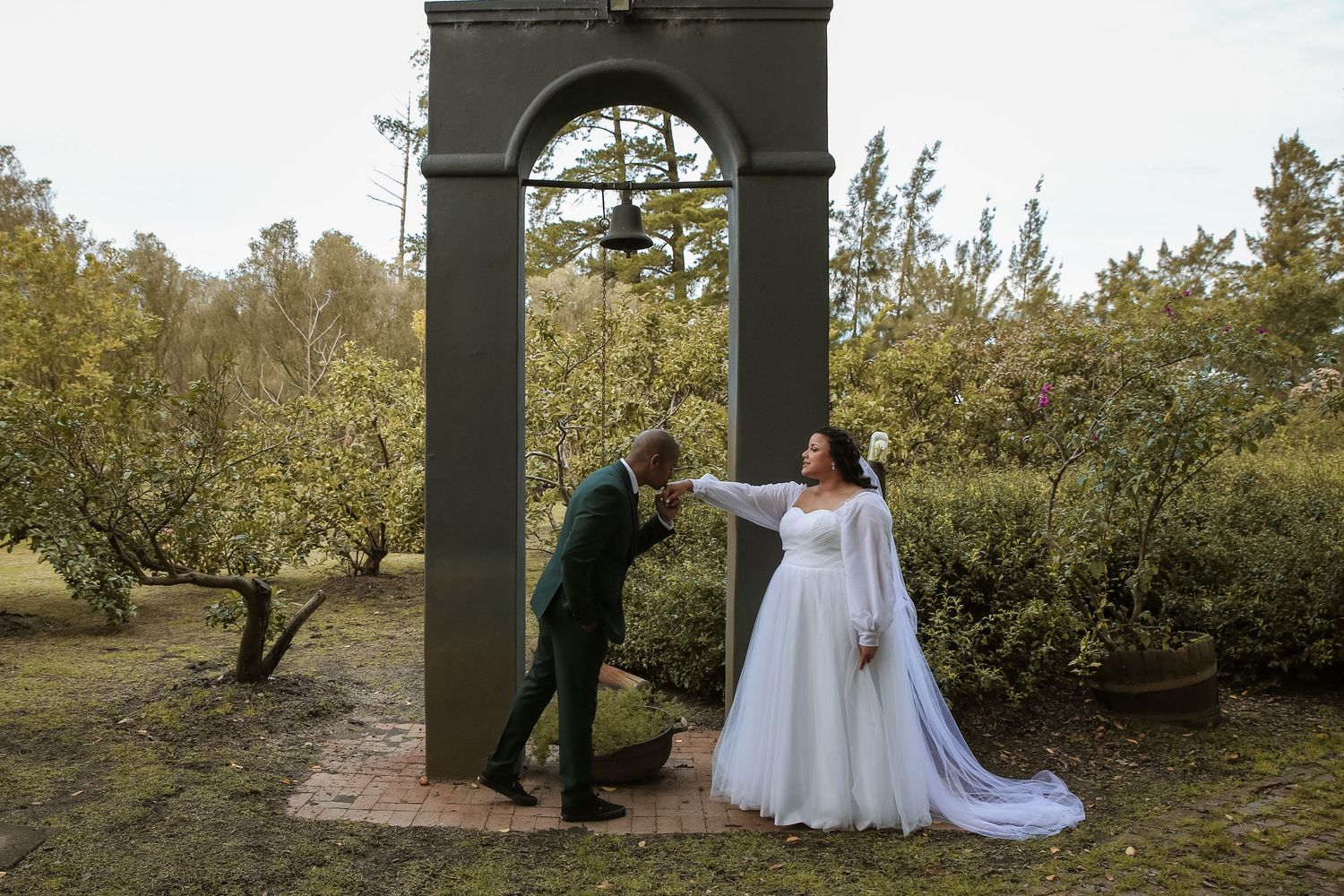Groom kissing bride's hand under stone arch in lush garden during outdoor wedding ceremony.