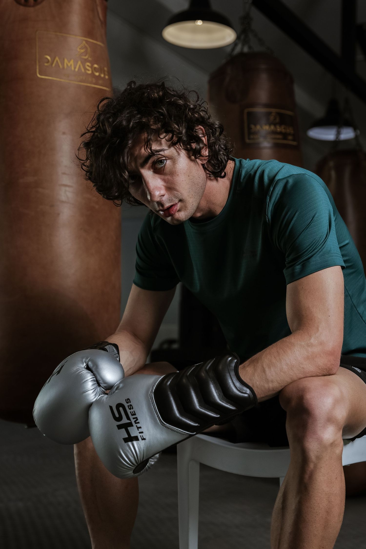 Male boxer in green shirt sits with silver boxing gloves beside a brown leather punching bag in a gym.