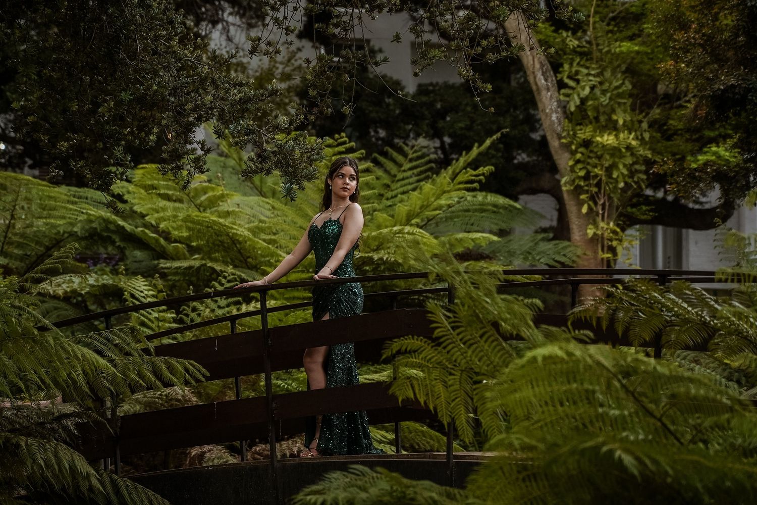 Woman in green sequin gown poses on wooden bridge surrounded by lush ferns in a garden.