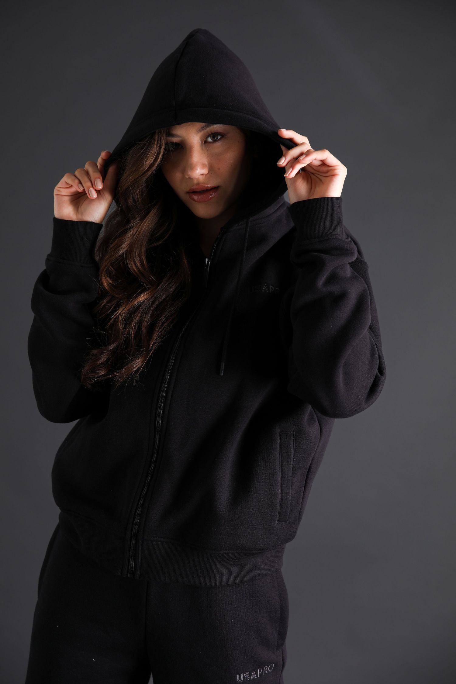 Woman in black zip-up hoodie and matching tracksuit pulls hood up in dark studio setting.