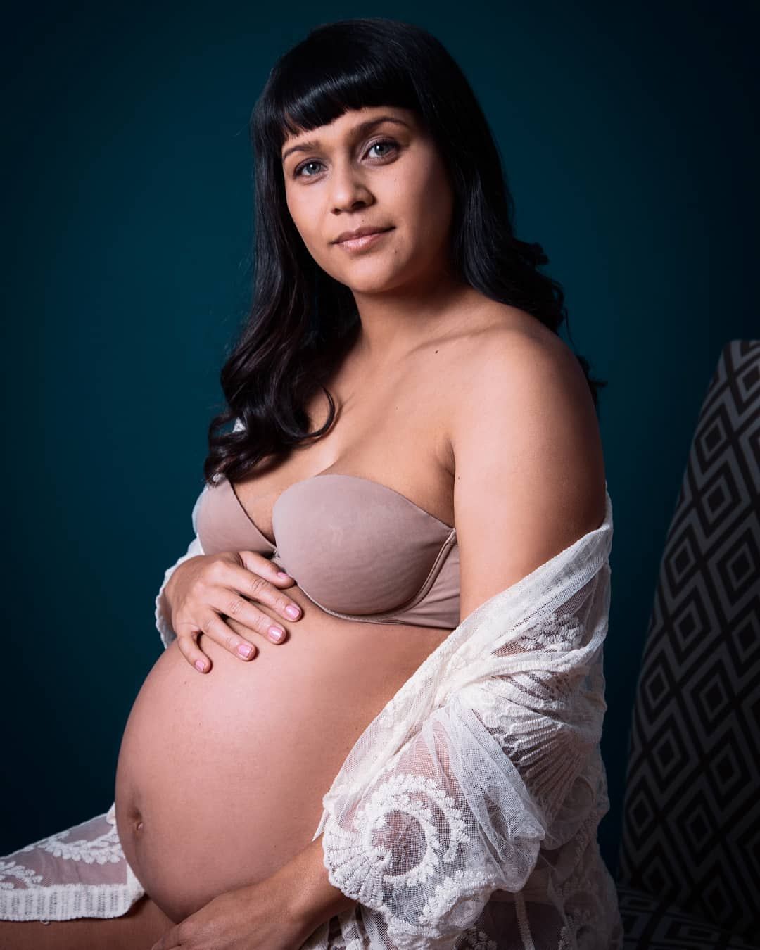 Pregnant woman in nude strapless bra and white lace robe holds baby bump against teal studio background.