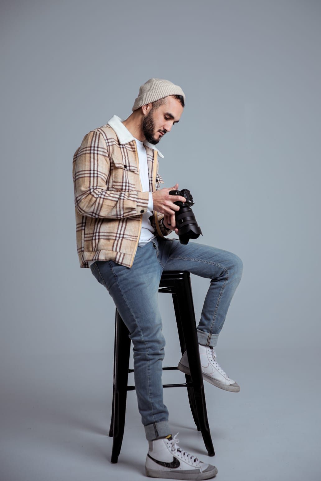 A bearded man in a plaid jacket and beanie sits on a stool, reviewing photos on a camera against a gray background.