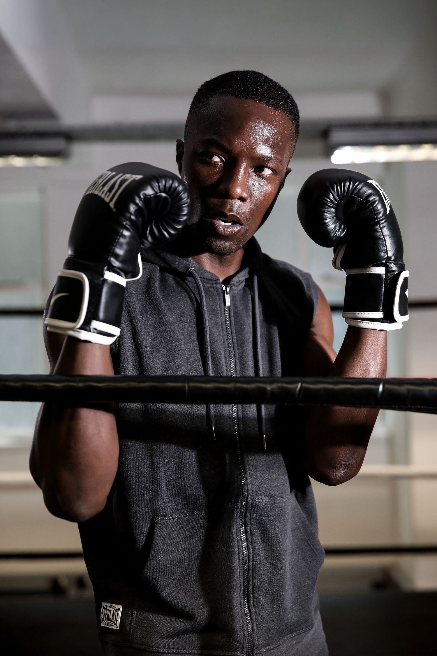 Male boxer in gray hoodie stands at boxing ring ropes wearing black boxing gloves in fighting stance.