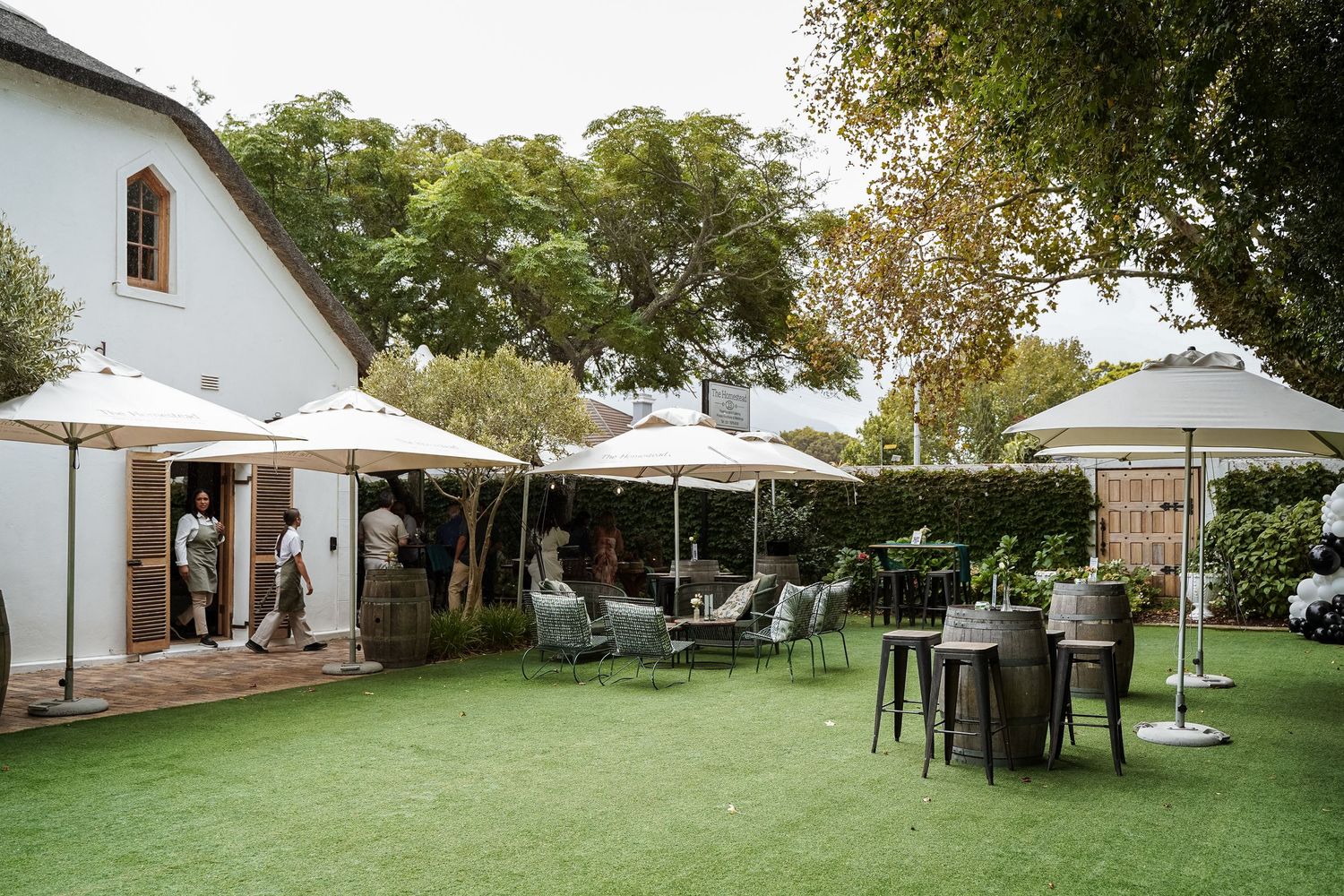 Outdoor garden event venue with white umbrellas, green lawn, and guests mingling near a white building.