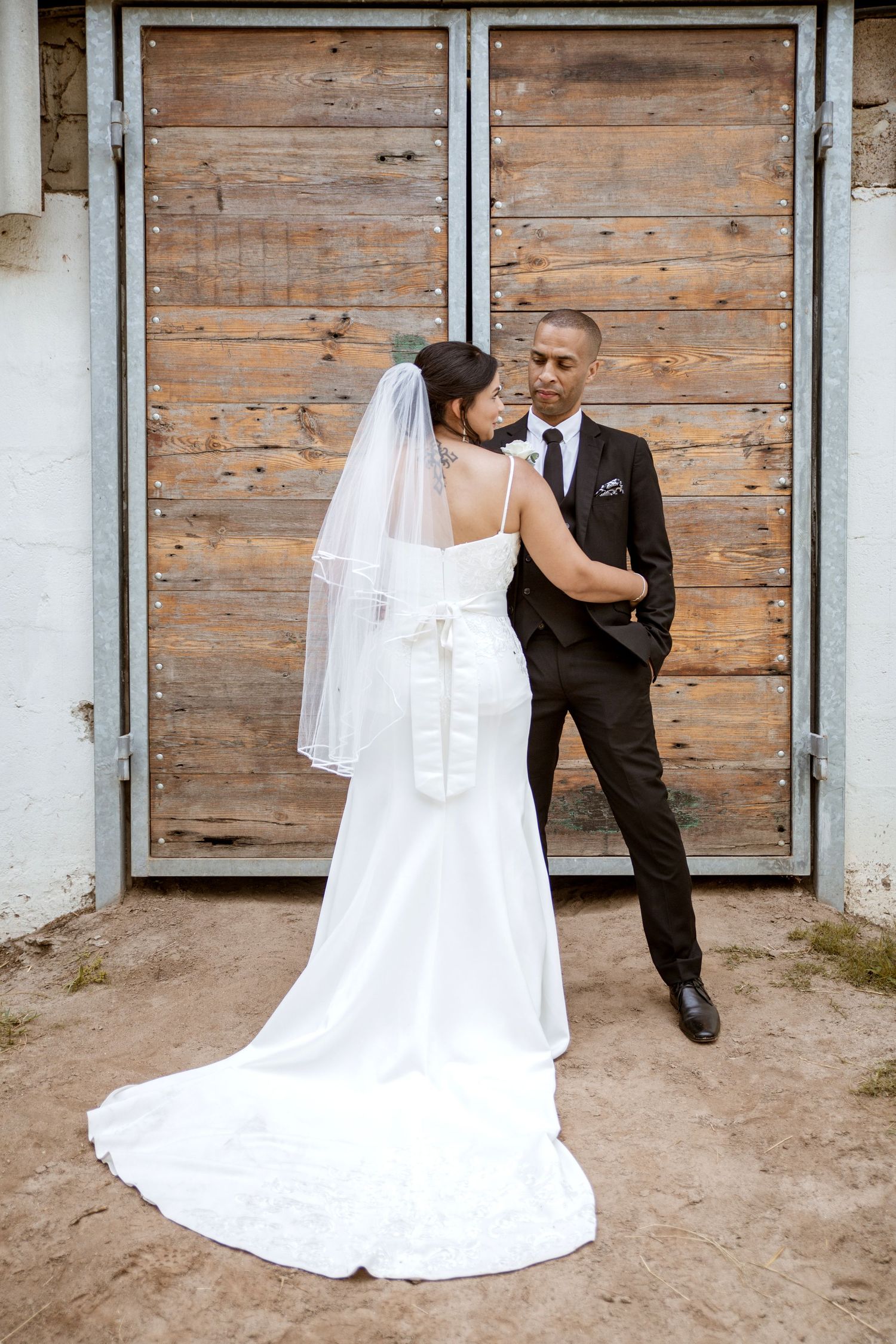 Bride in white gown with veil and groom in black suit posing by wooden doors.