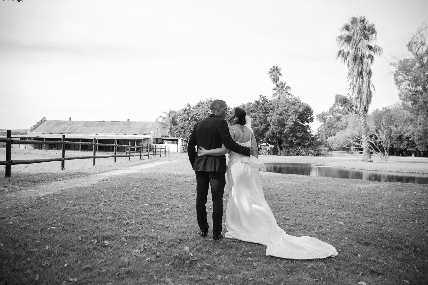Black and white photo of bride and groom walking hand in hand toward a pond at an outdoor wedding venue.