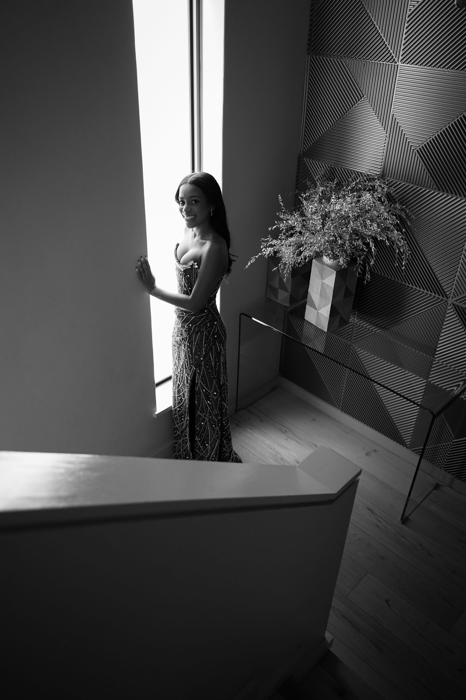 Elegant woman in sequin gown standing by tall window on staircase landing, black and white photo.
