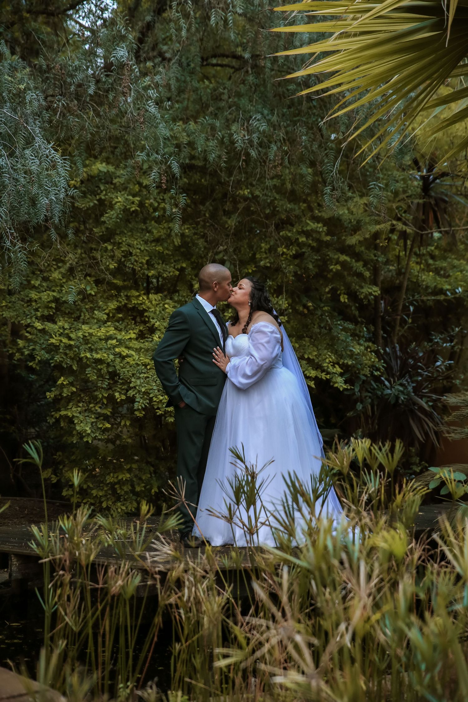 Bride and groom sharing romantic kiss surrounded by lush green trees beside tranquil garden pond.