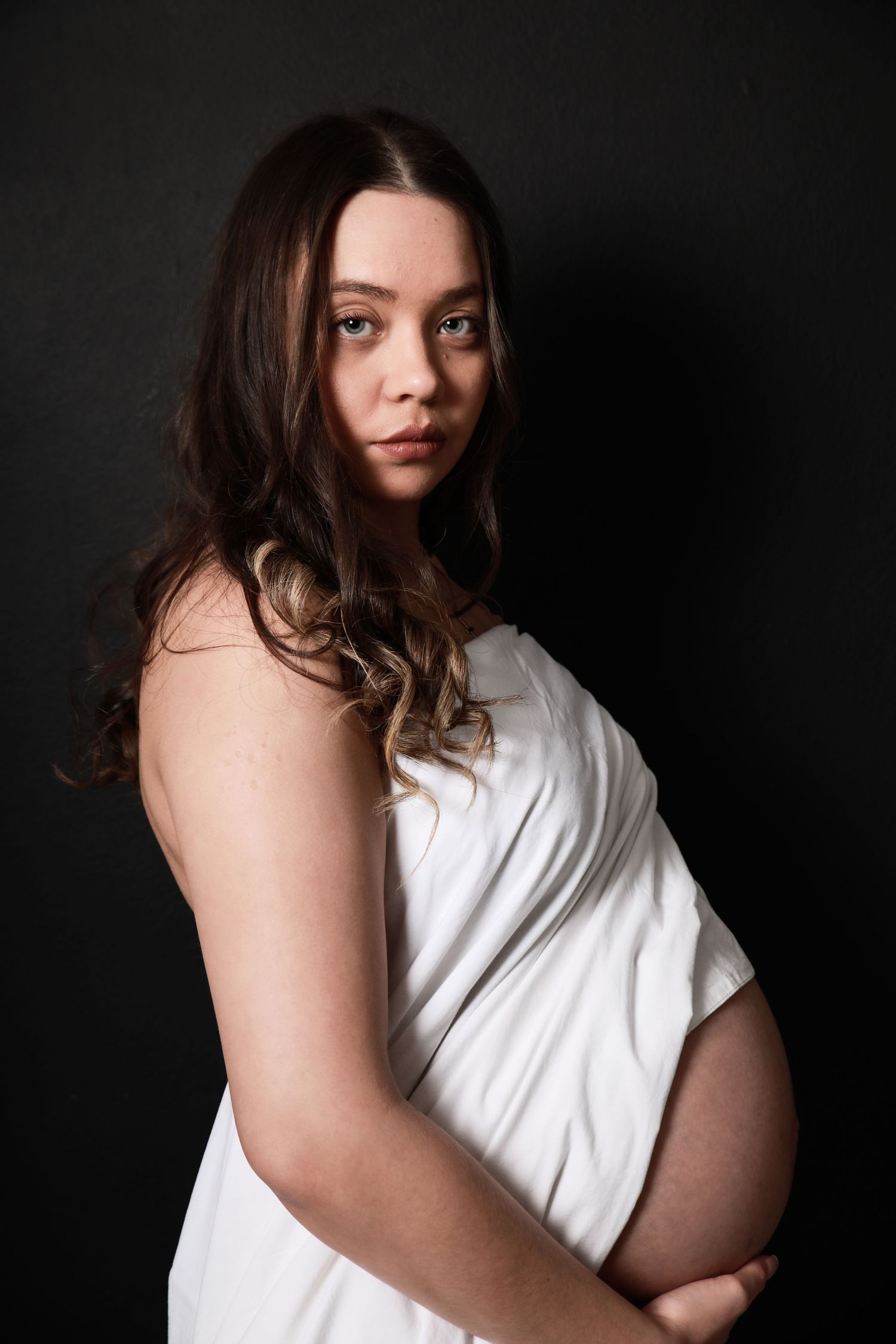 Pregnant woman wrapped in white fabric gazes at camera in moody dark studio maternity portrait.