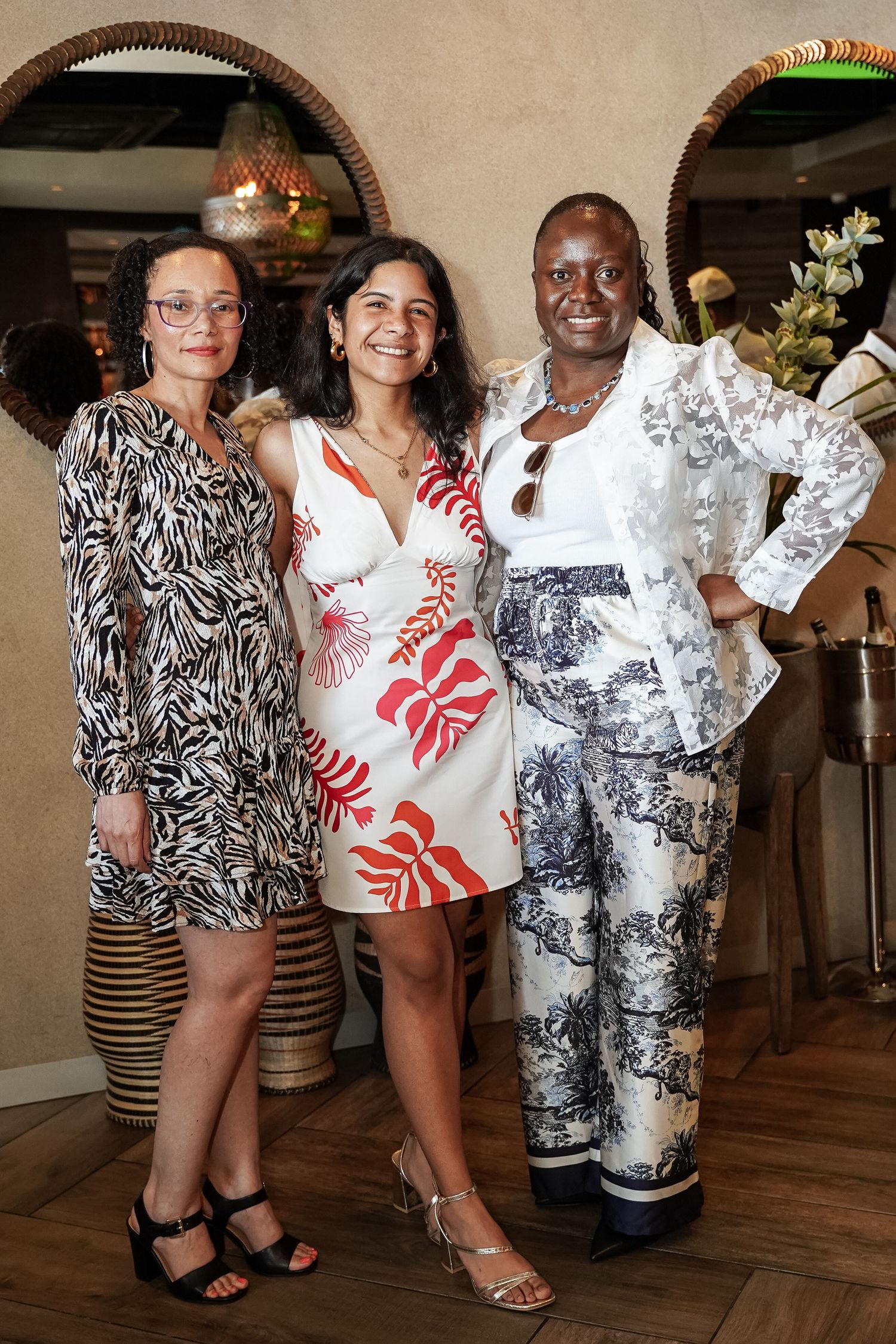 Three women posing together in stylish patterned outfits at decorated interior venue.