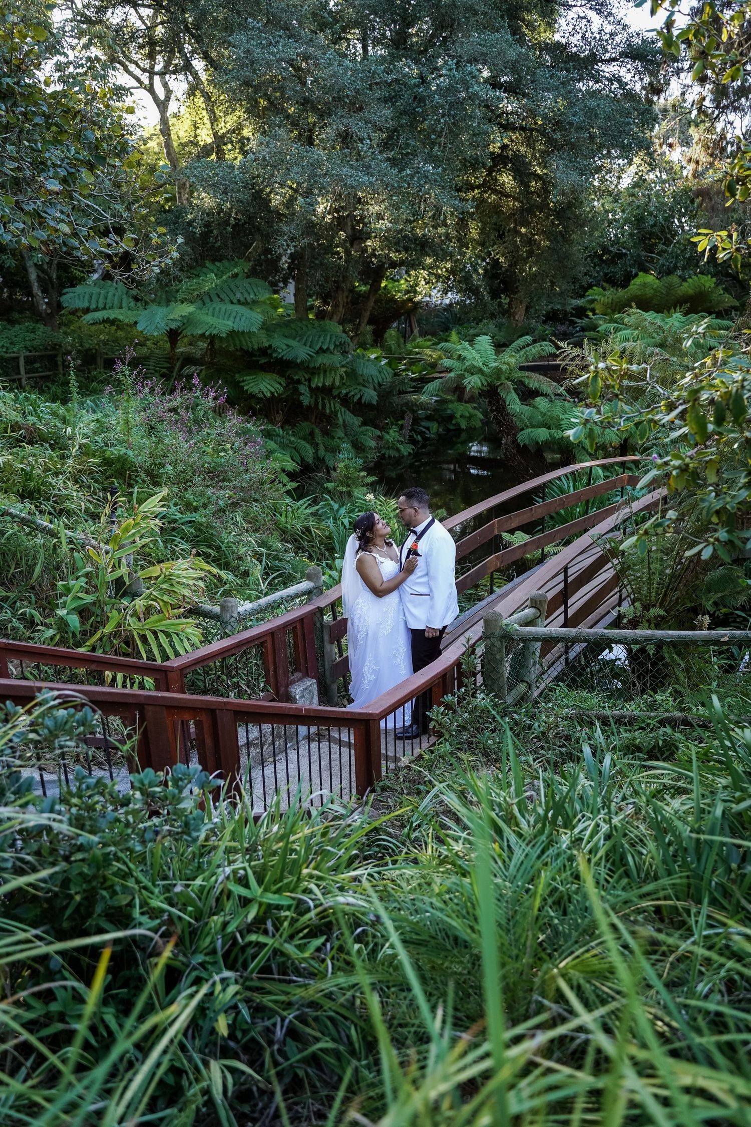 Bride and groom embrace on a wooden bridge surrounded by lush tropical greenery during an outdoor wedding photoshoot.