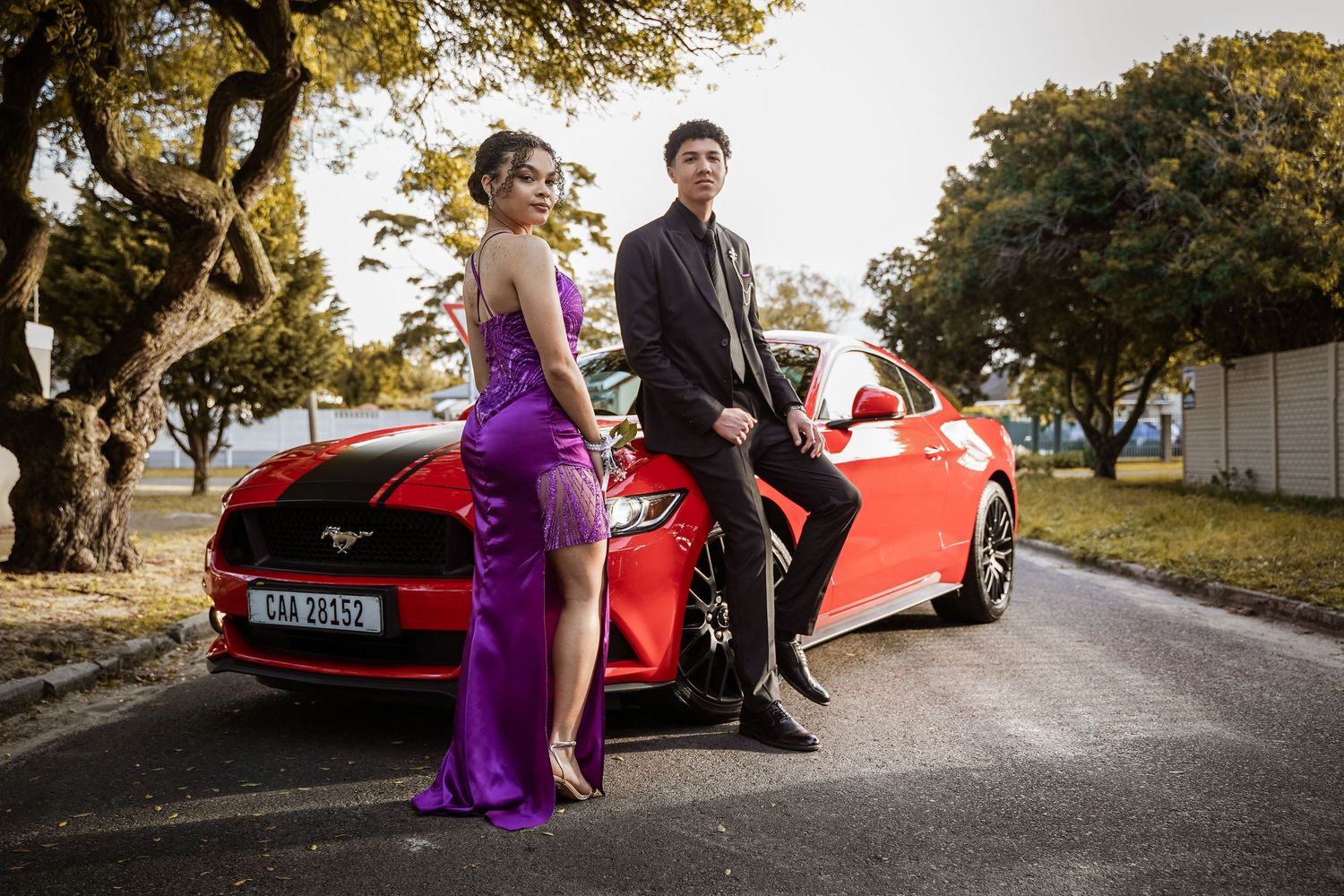 Elegant couple in formal wear posing beside a red Ford Mustang GT.