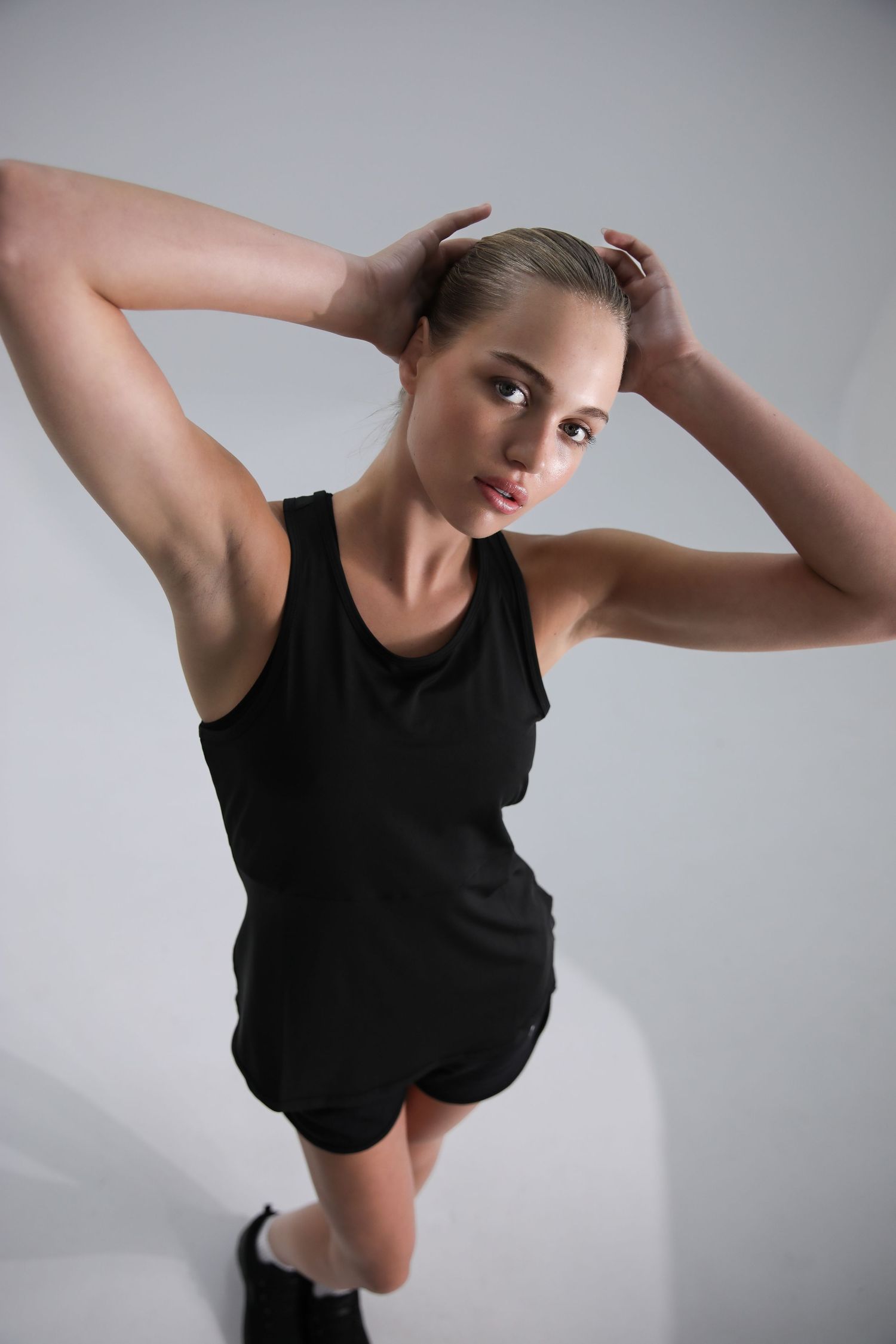 Fit woman in black tank top and shorts posing with arms raised on light background.