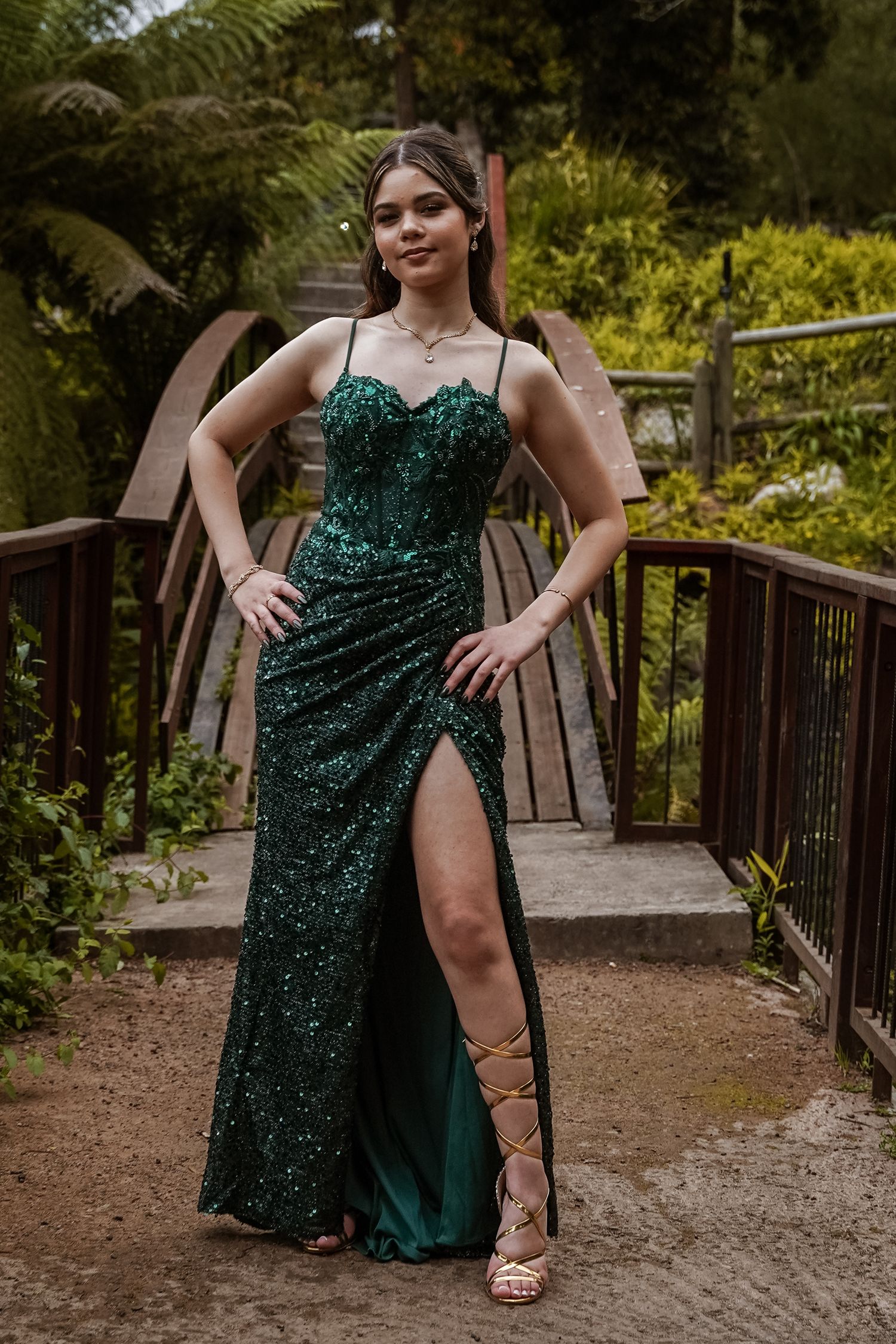 Young woman in green sequin formal gown with high slit and strappy heels poses outdoors on bridge.