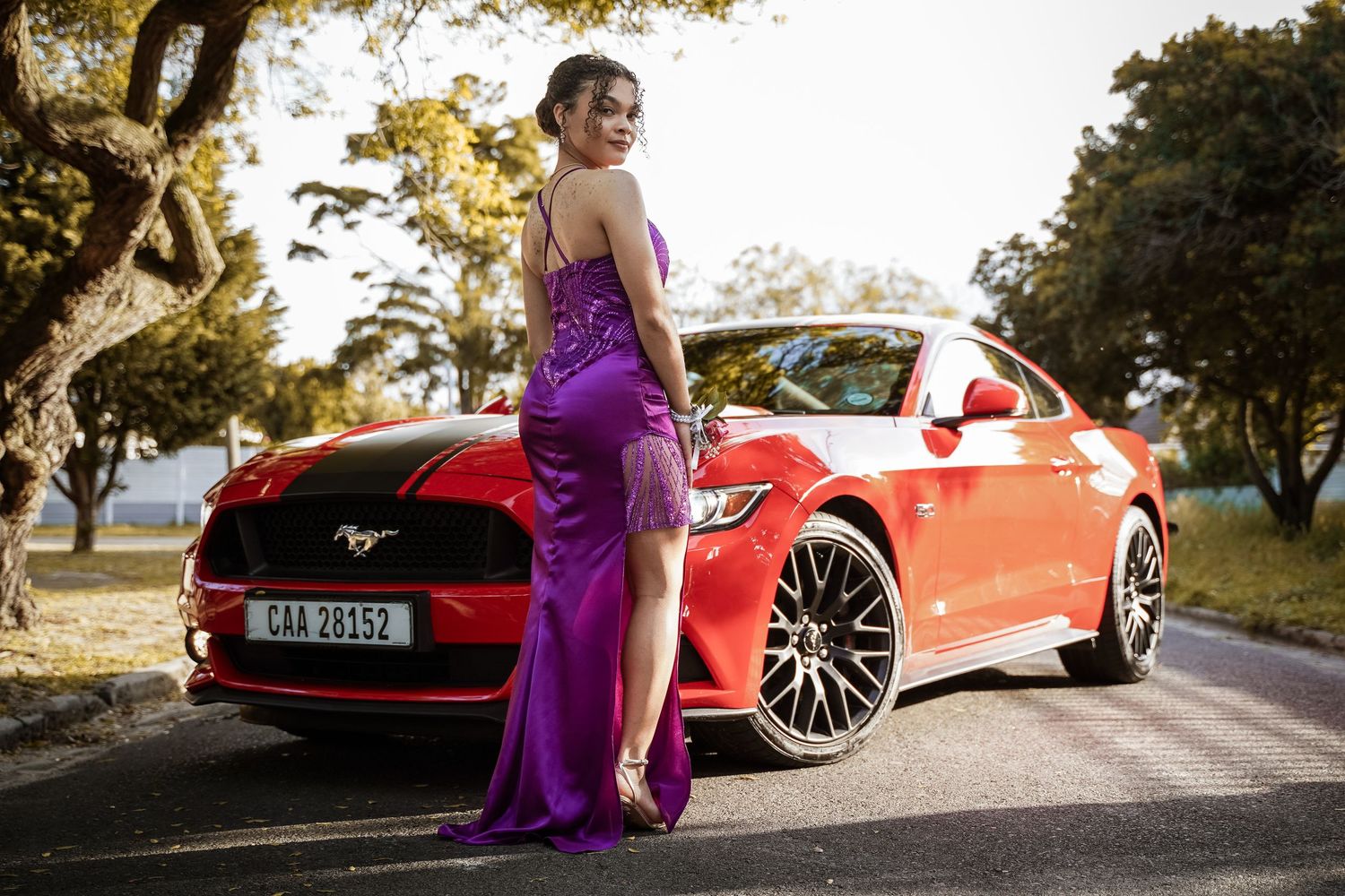 Young woman in purple sequin gown posing beside red Ford Mustang on tree-lined road at sunset.