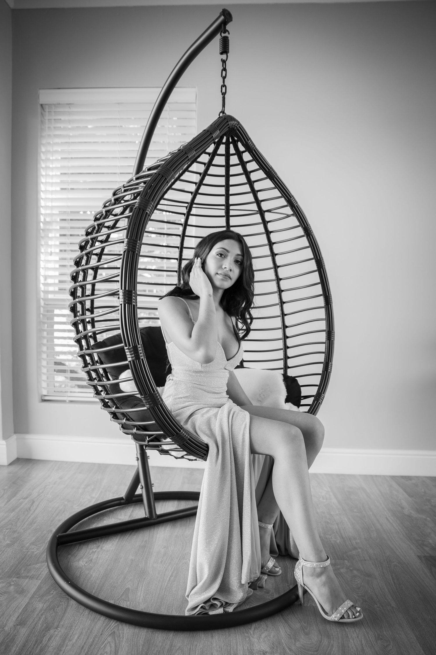 Woman in elegant white dress seated in hanging rattan egg chair, black and white portrait.