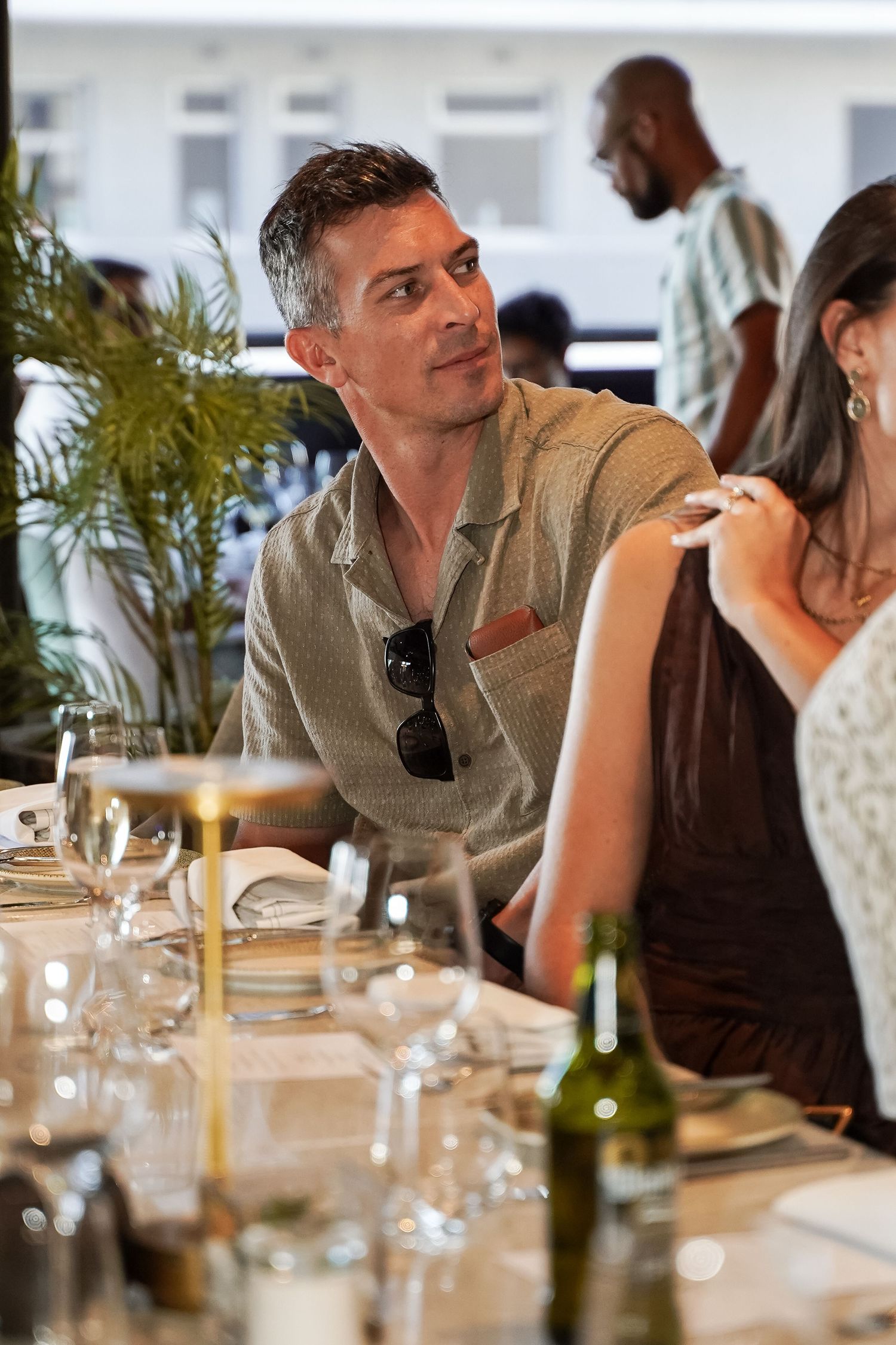 Man in grey linen shirt with sunglasses sitting at elegant dinner table setting.