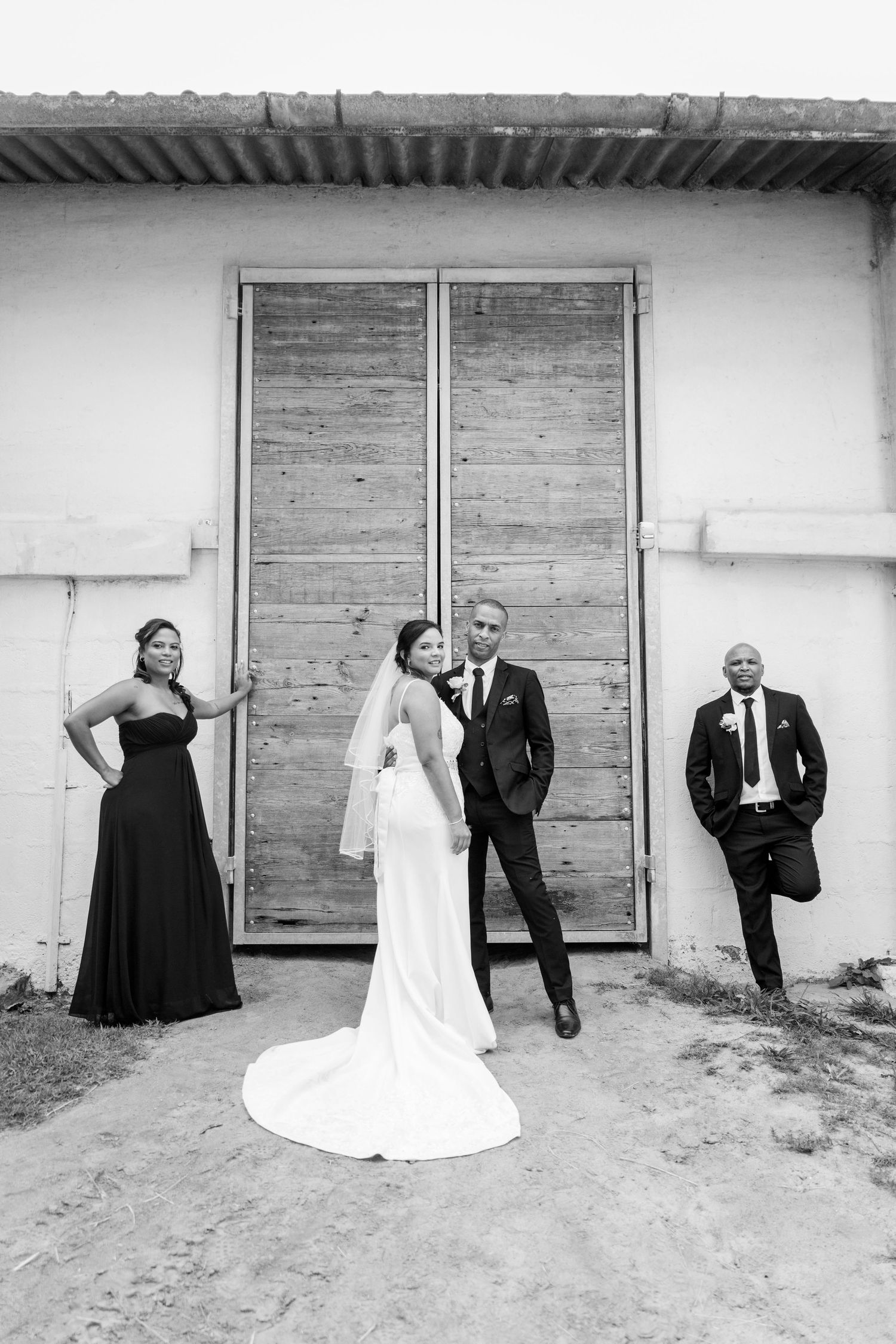 Black and white wedding photo of bride, groom, bridesmaid, and groomsman posing before rustic wooden doors.