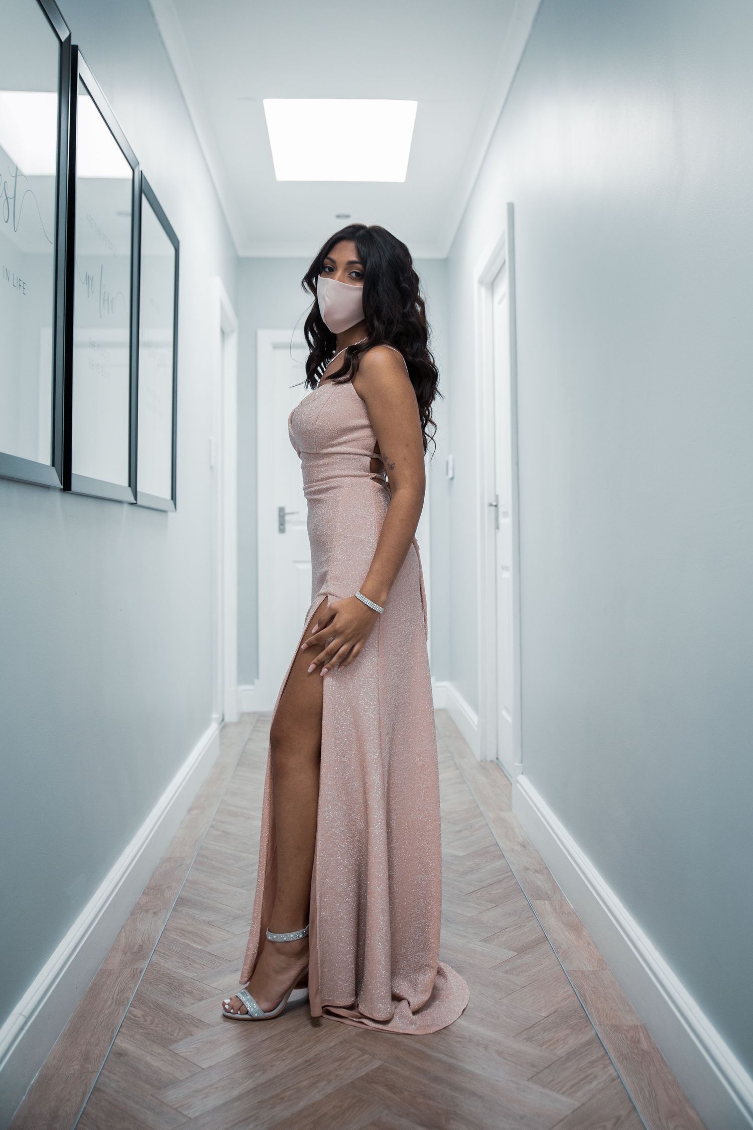 Woman in blush pink strapless gown wearing face mask posing in modern white hallway.