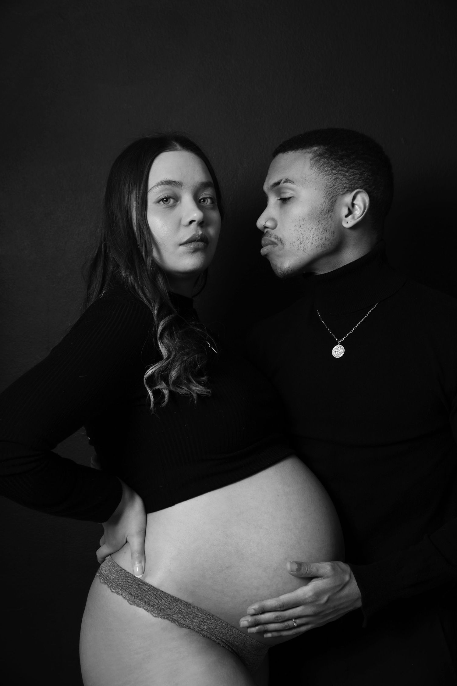 Black and white portrait of couple, man kissing pregnant womans shoulder while holding her bump.