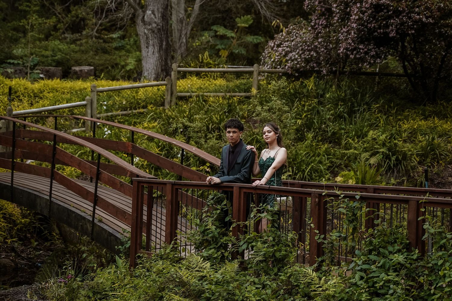 Couple in formal attire pose together on a winding wooden bridge surrounded by lush greenery.