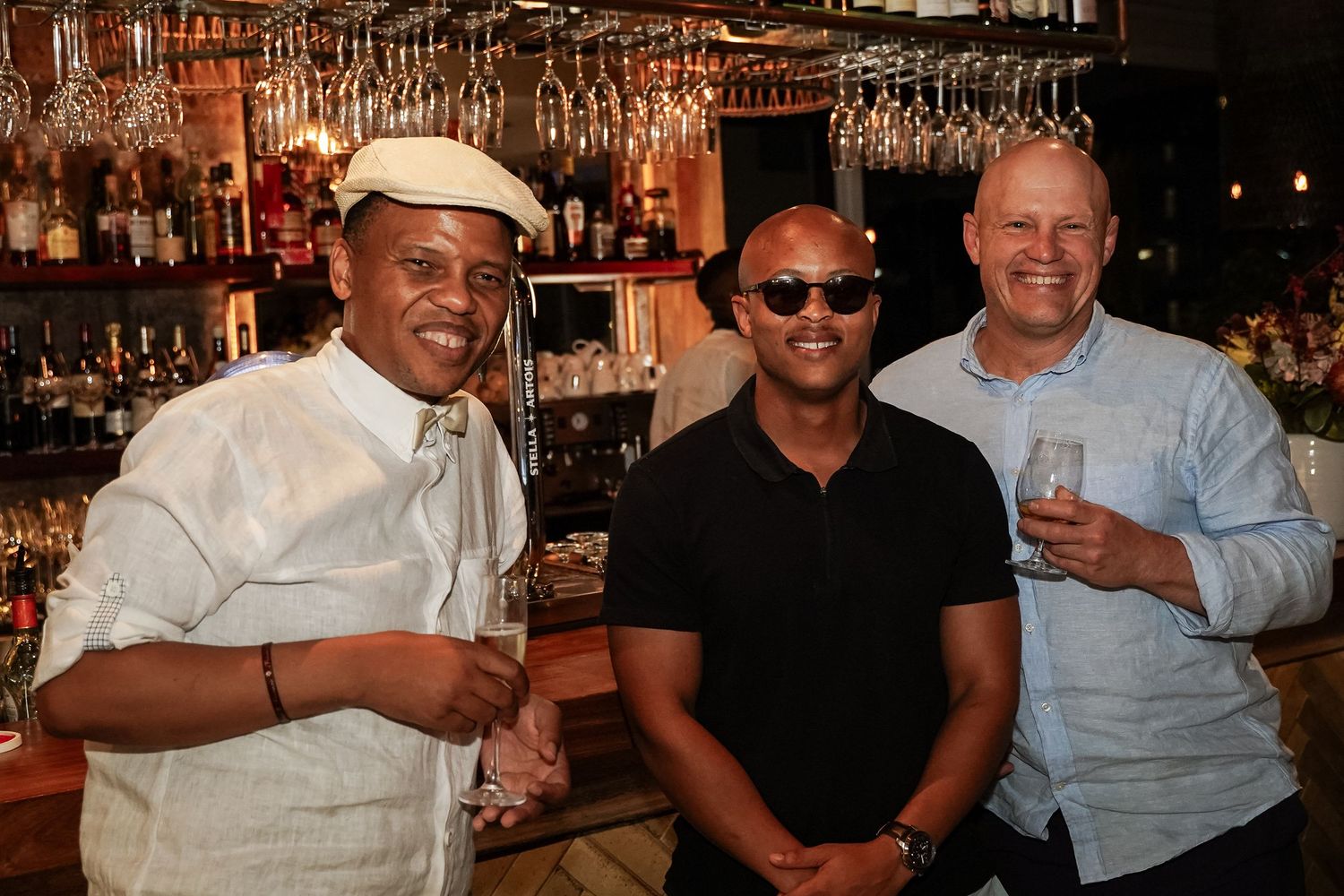 Three men smiling together at bar with hanging glassware in background.