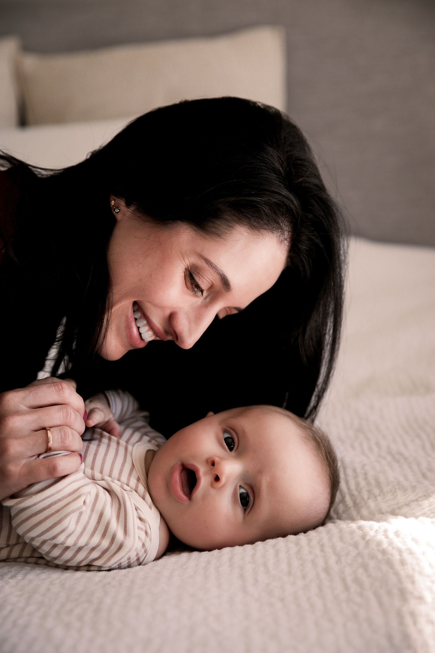 Dark-haired mother smiling warmly at her baby lying on a white textured bed.