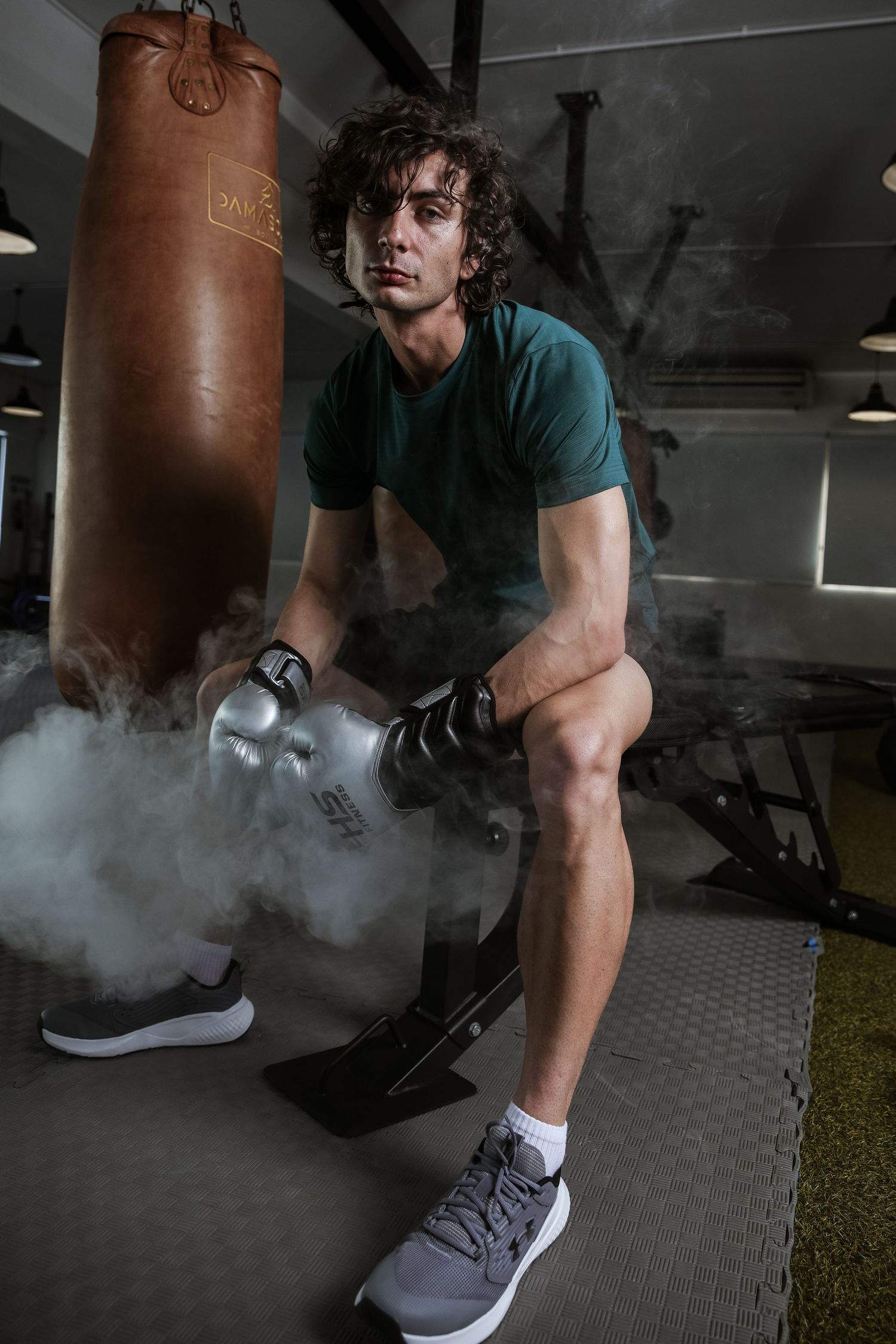 Athlete in green shirt sits with boxing gloves amid dramatic smoke near a punching bag in a dark gym.