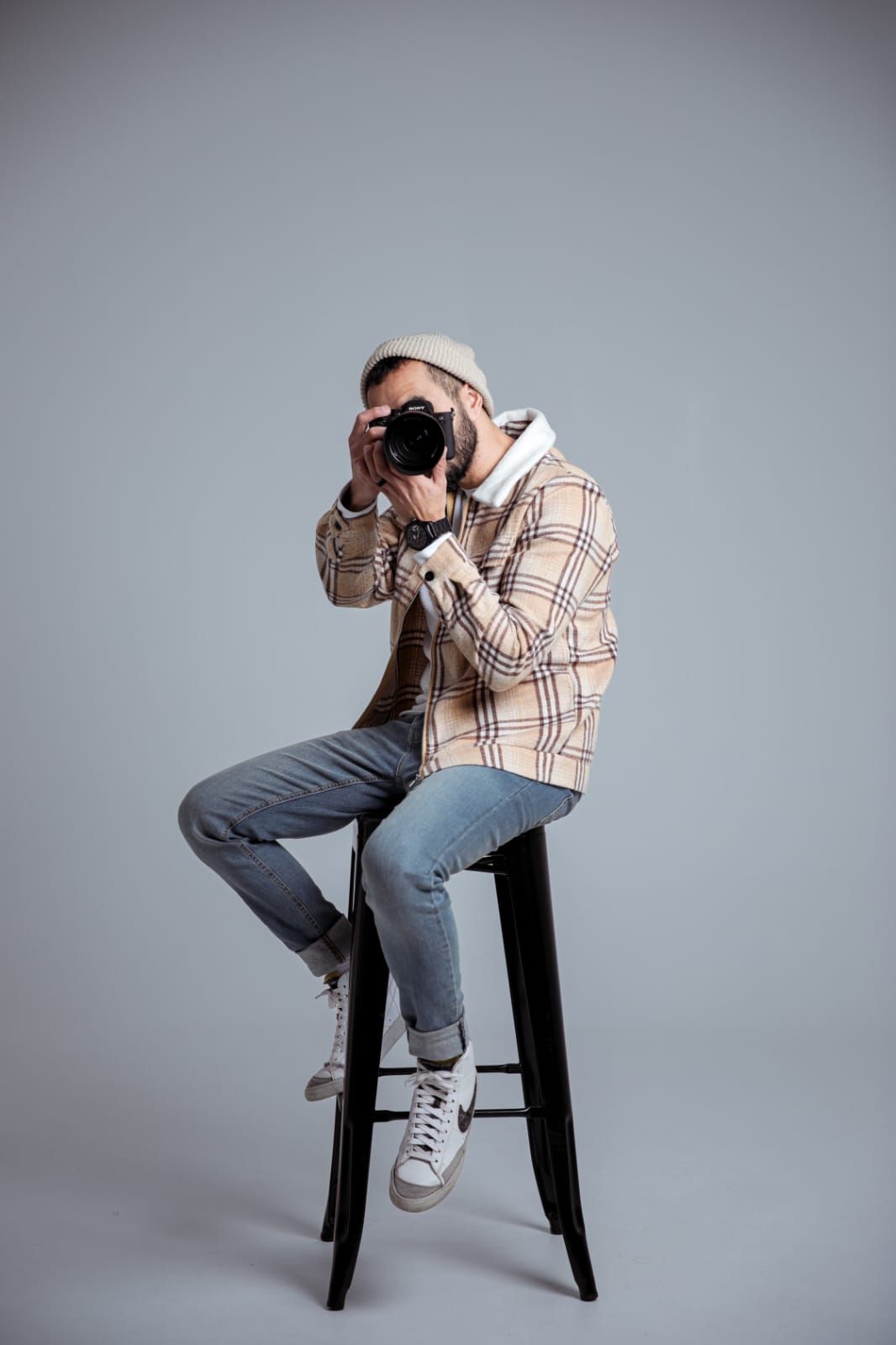 Man in plaid shirt and cap sitting on stool, taking photo with DSLR camera against gray background.