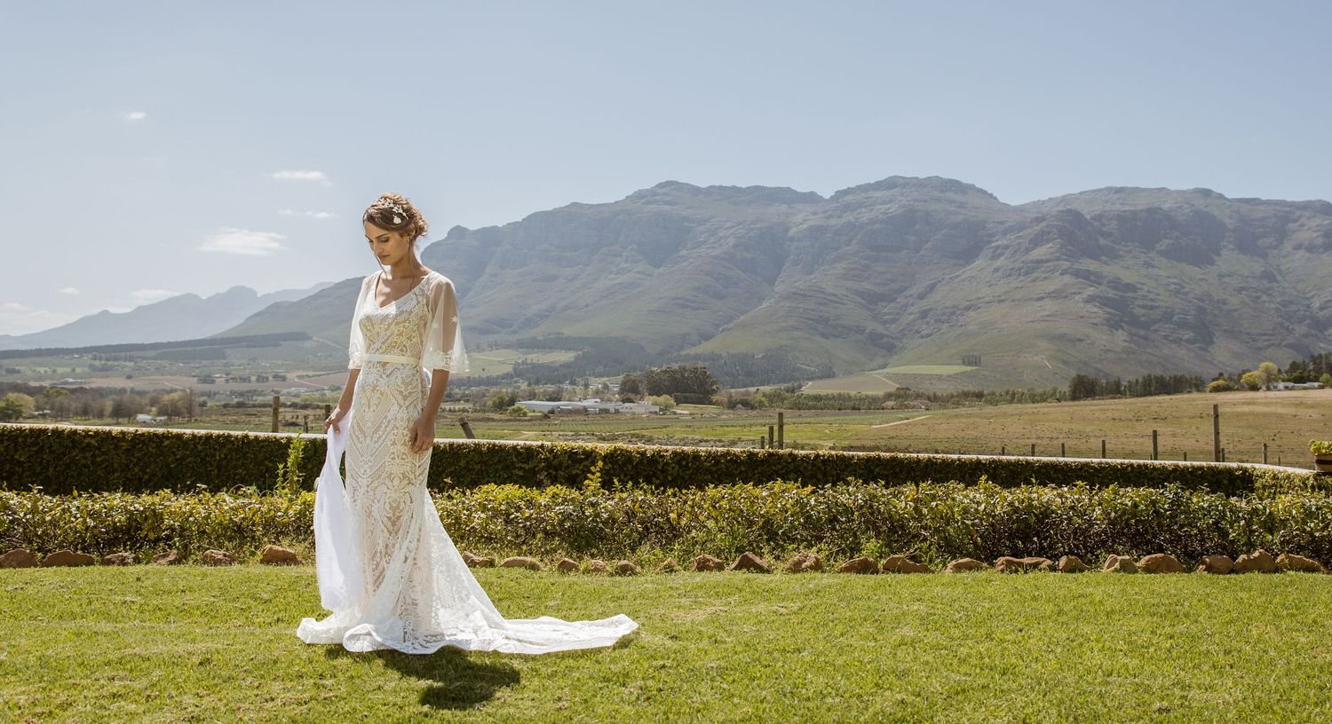 Bride in white lace wedding gown with train stands on green lawn with scenic mountain backdrop on sunny day.