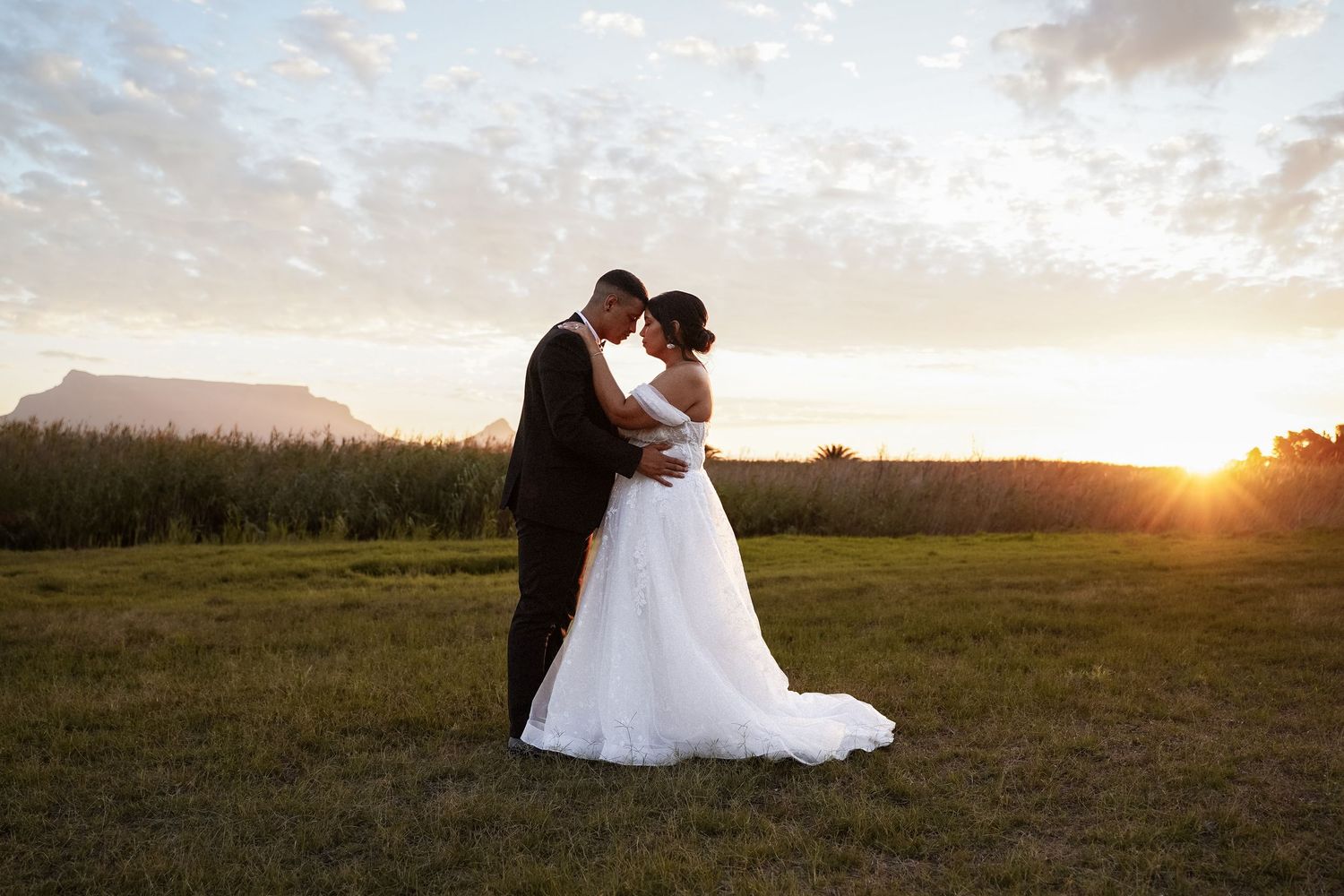 Bride and groom embracing at sunset in a green field with Table Mountain silhouette in the background.