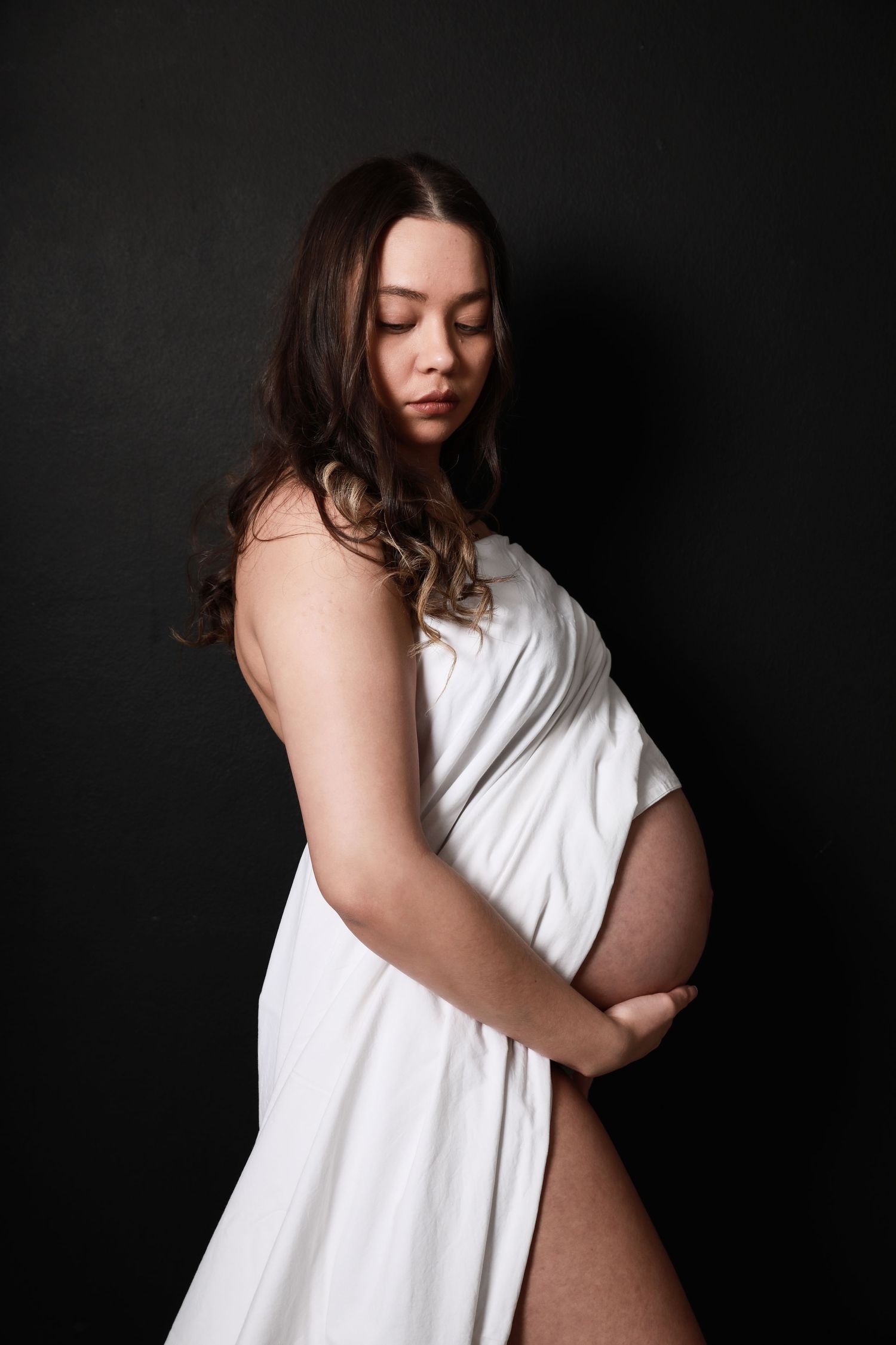 Pregnant woman draped in white fabric holds baby bump while looking down on black background.