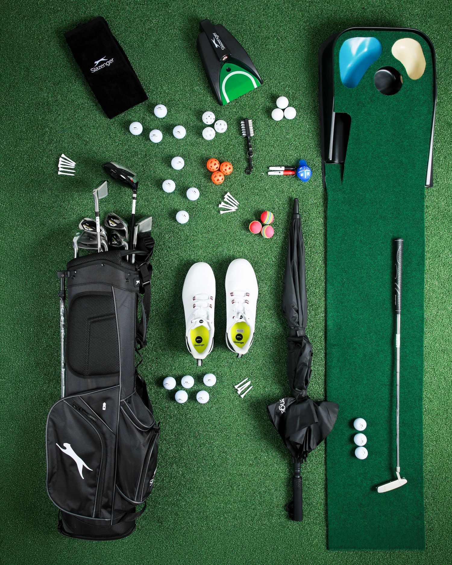 Flat lay of golf equipment including clubs, bag, balls, shoes, umbrella and putting mat on green turf.