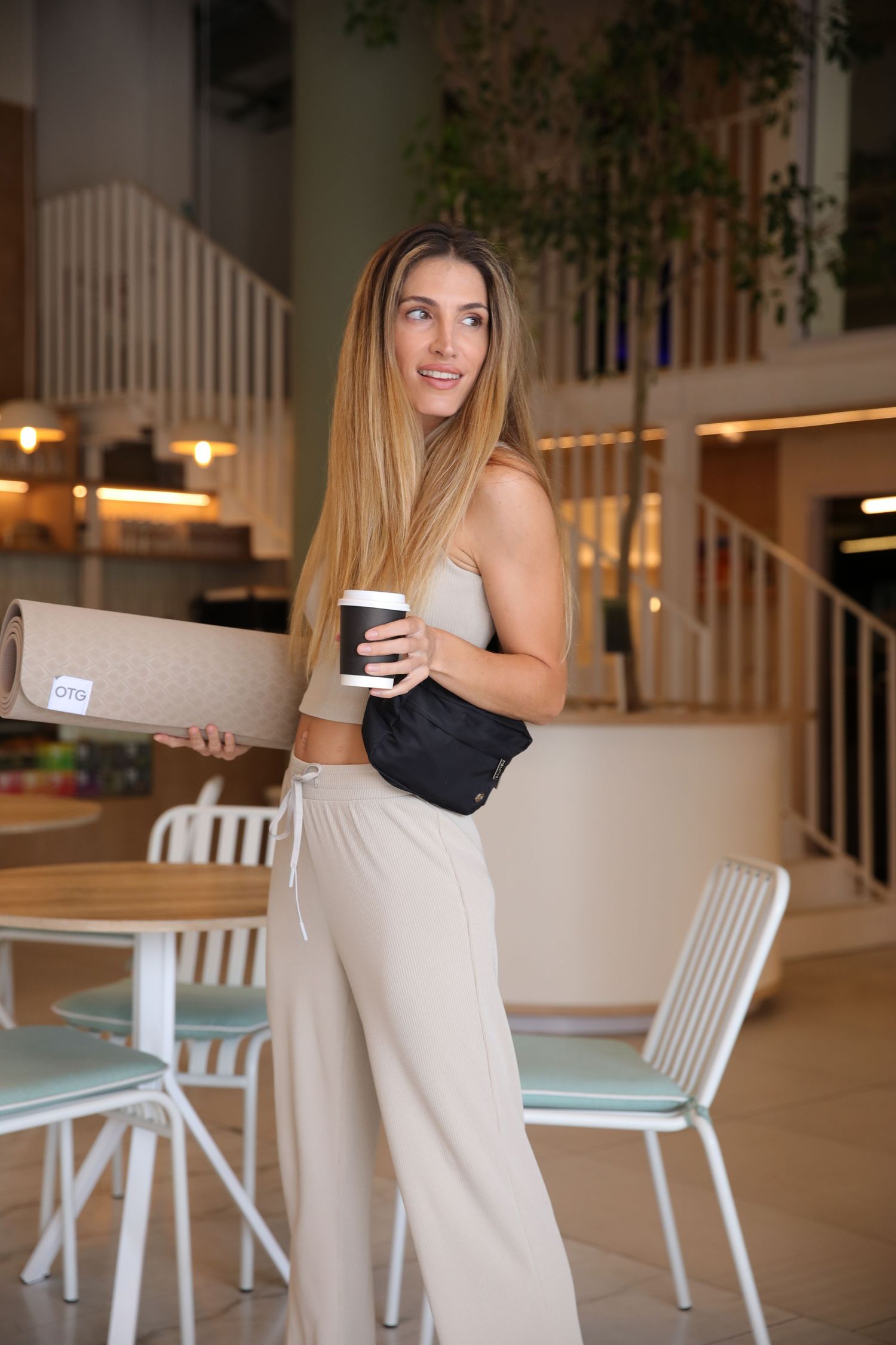 Fit woman in beige athleisure holding rolled yoga mat and coffee cup in modern space.