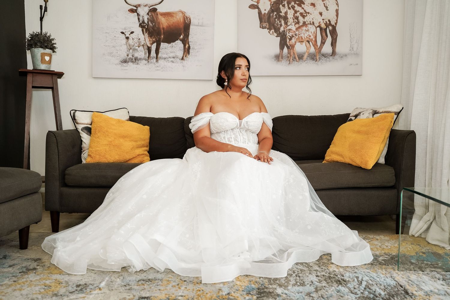 Bride in off-shoulder ballgown wedding dress sitting gracefully on sofa with floral artwork.