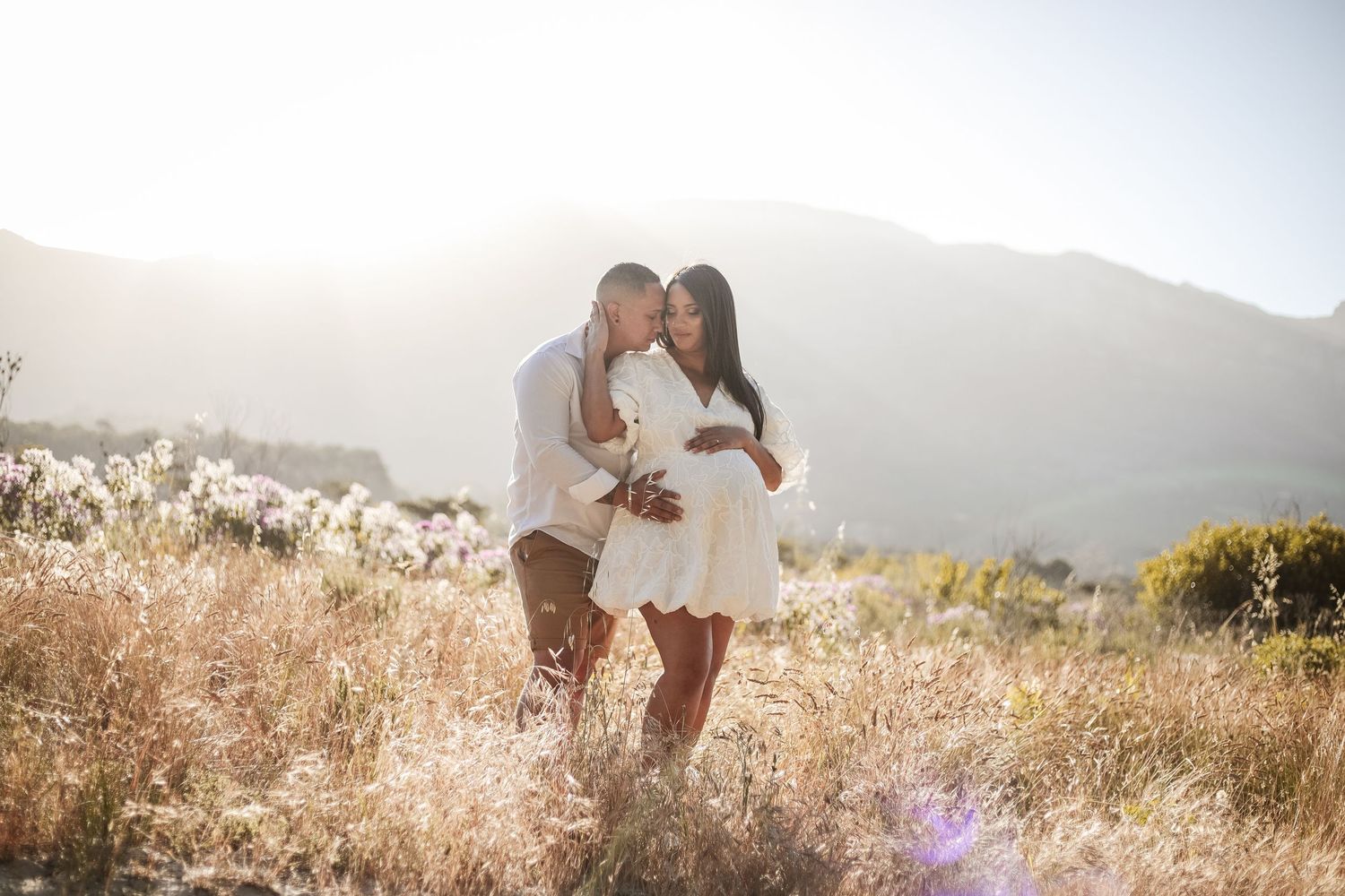 Couple shares tender kiss in sunlit field with mountain backdrop during outdoor maternity photoshoot.