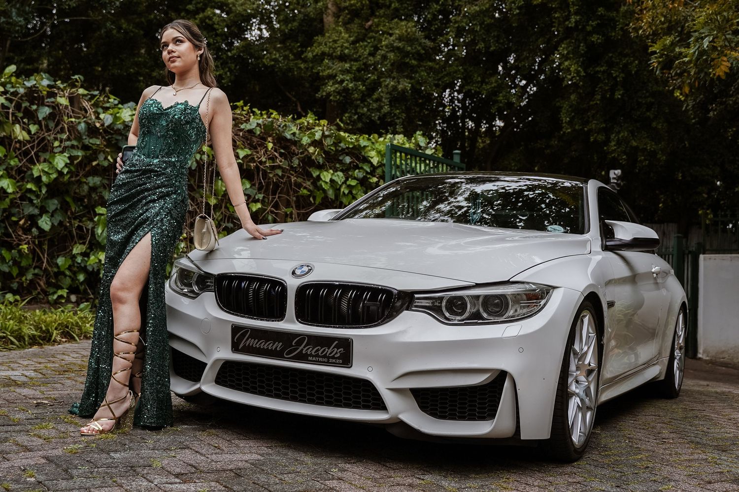 Woman in green sequin slit gown poses beside white BMW M4 convertible in tree-lined driveway.