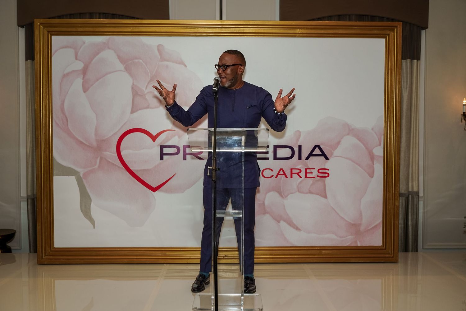 Man in navy suit speaking at a Primedia Cares event in front of a floral backdrop.