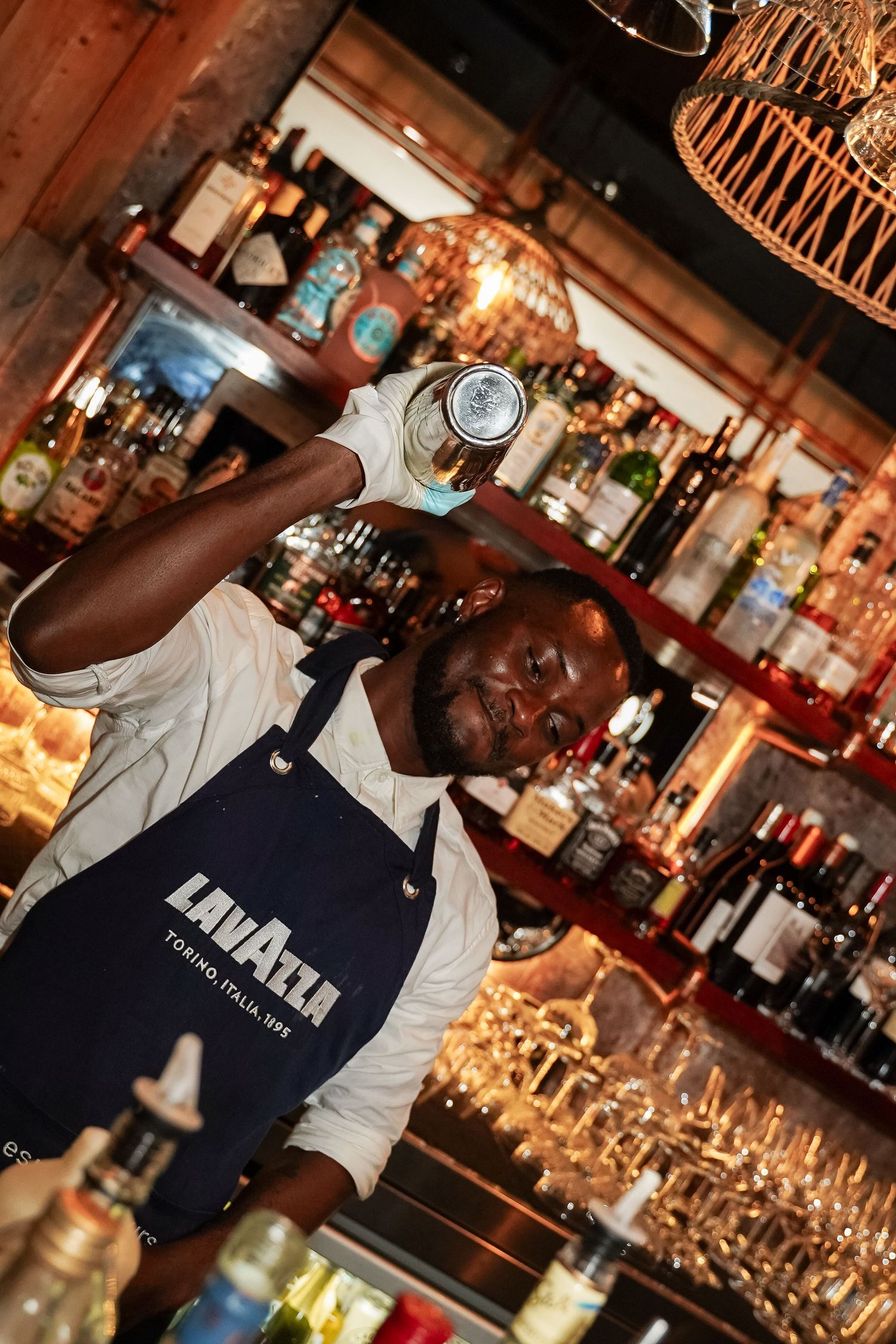 Bartender in Lavazza apron performing cocktail shaker flair behind fully stocked bar.