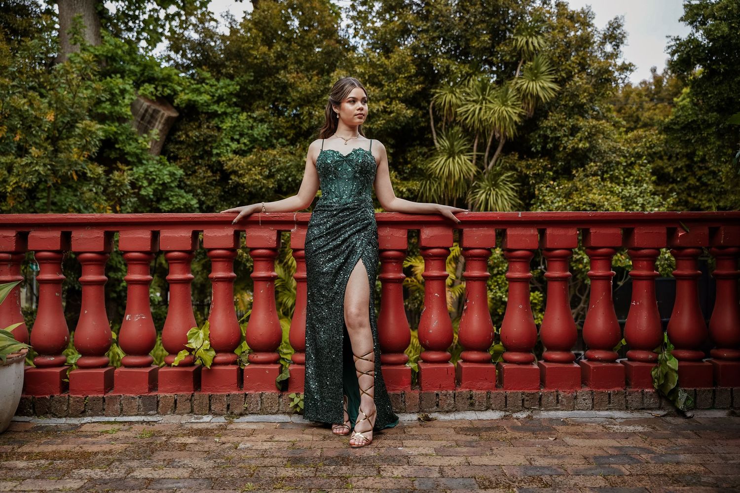 Woman in green sequin formal gown with high slit poses against red balustrade with garden backdrop.
