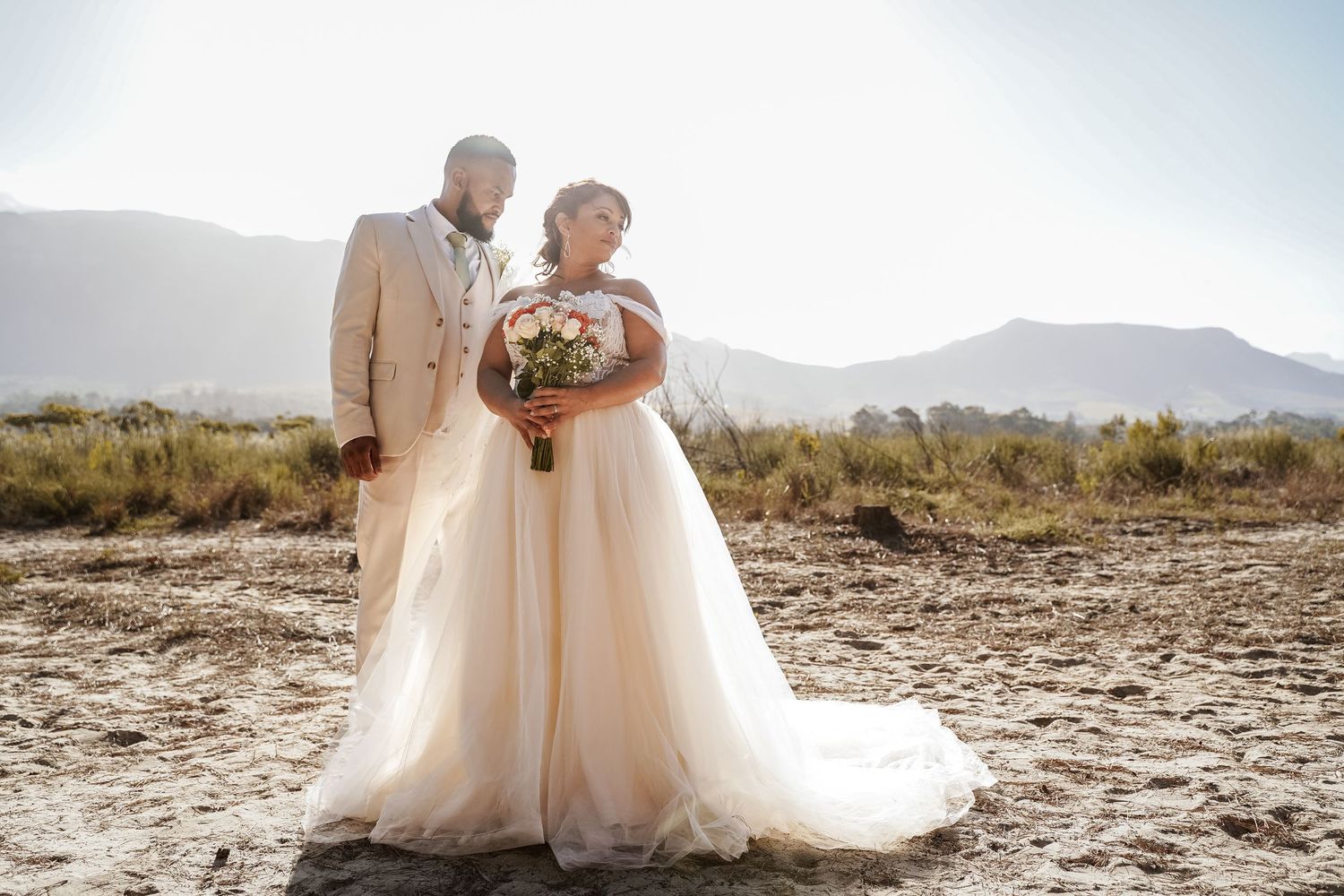Bride holding floral bouquet standing with bearded groom in tan suit against mountain backdrop.