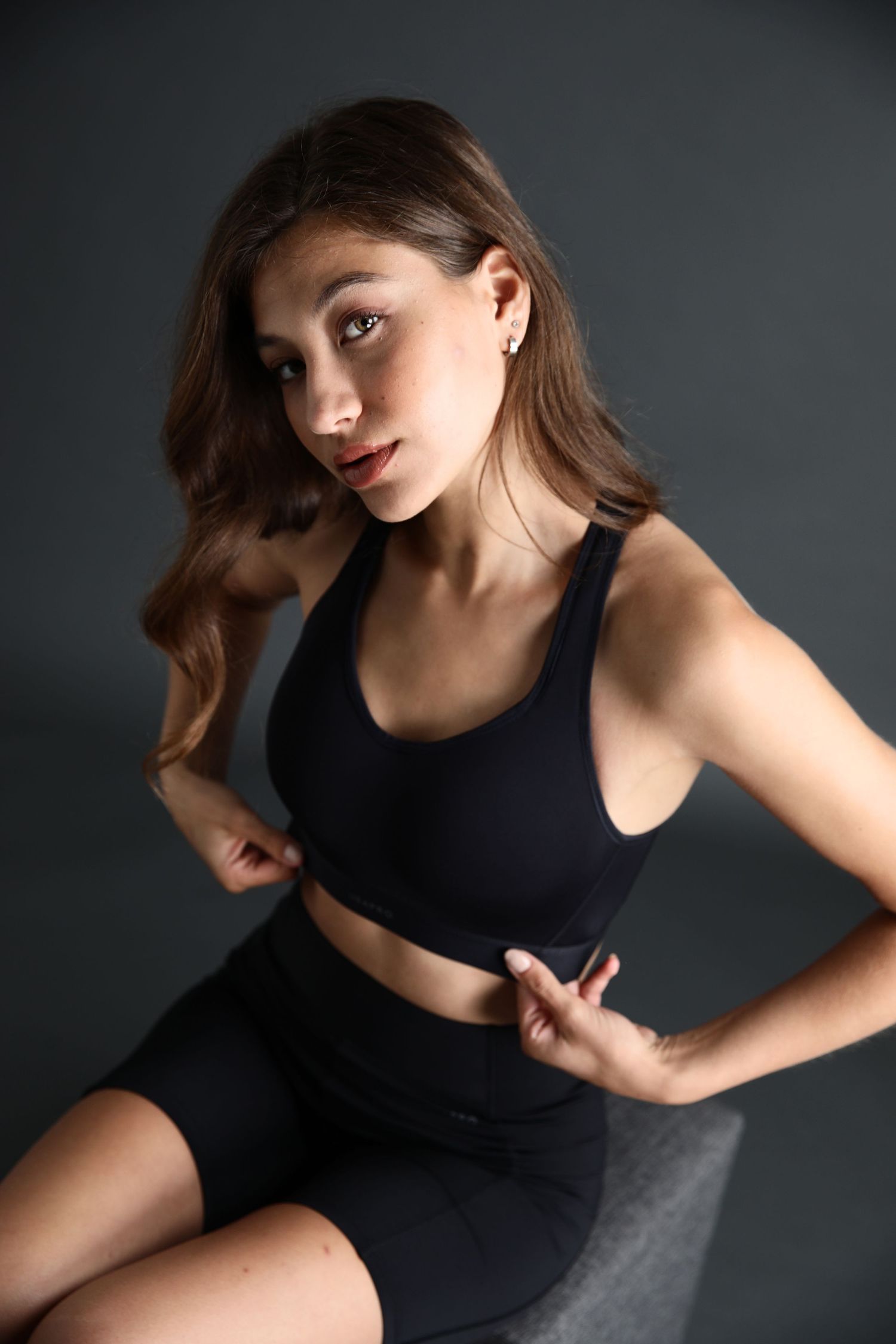 Woman in black sports bra and shorts sitting and posing on gray background.