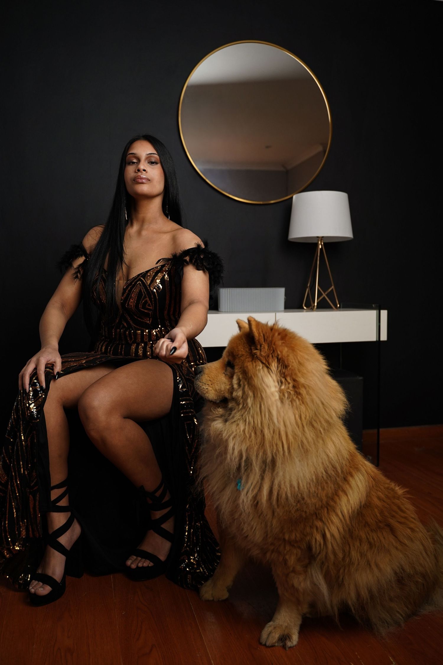 Woman in black gold sequin dress seated with a Chow Chow dog by a mirror.