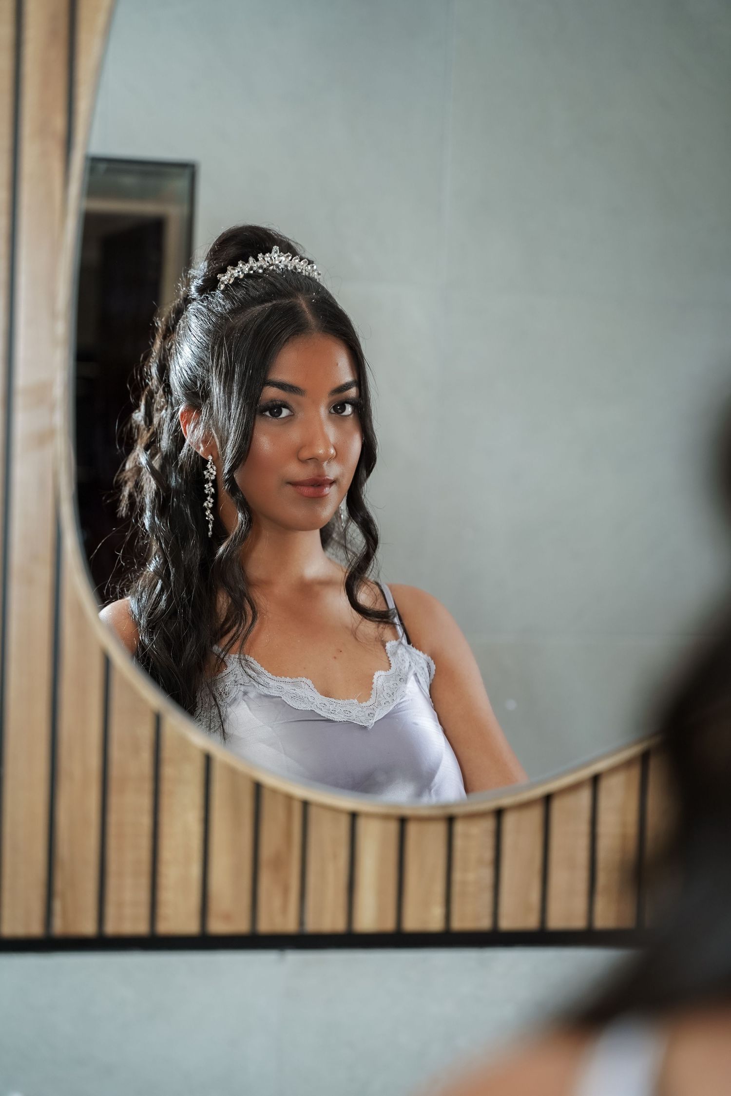 Elegant young woman with tiara and drop earrings reflected in ornate mirror wearing white lace dress.