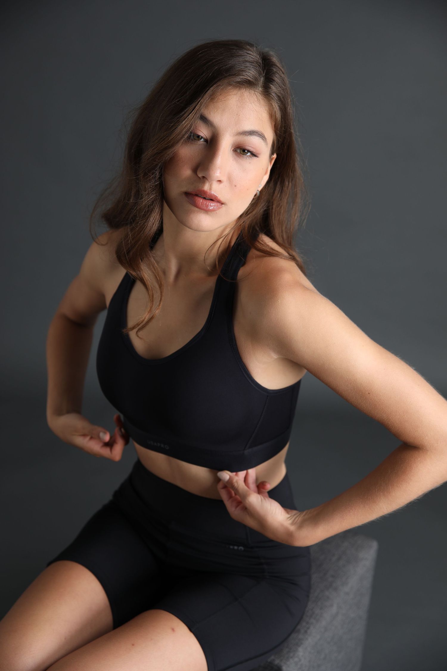 Woman in black sports bra and bike shorts sitting confidently on gray background.